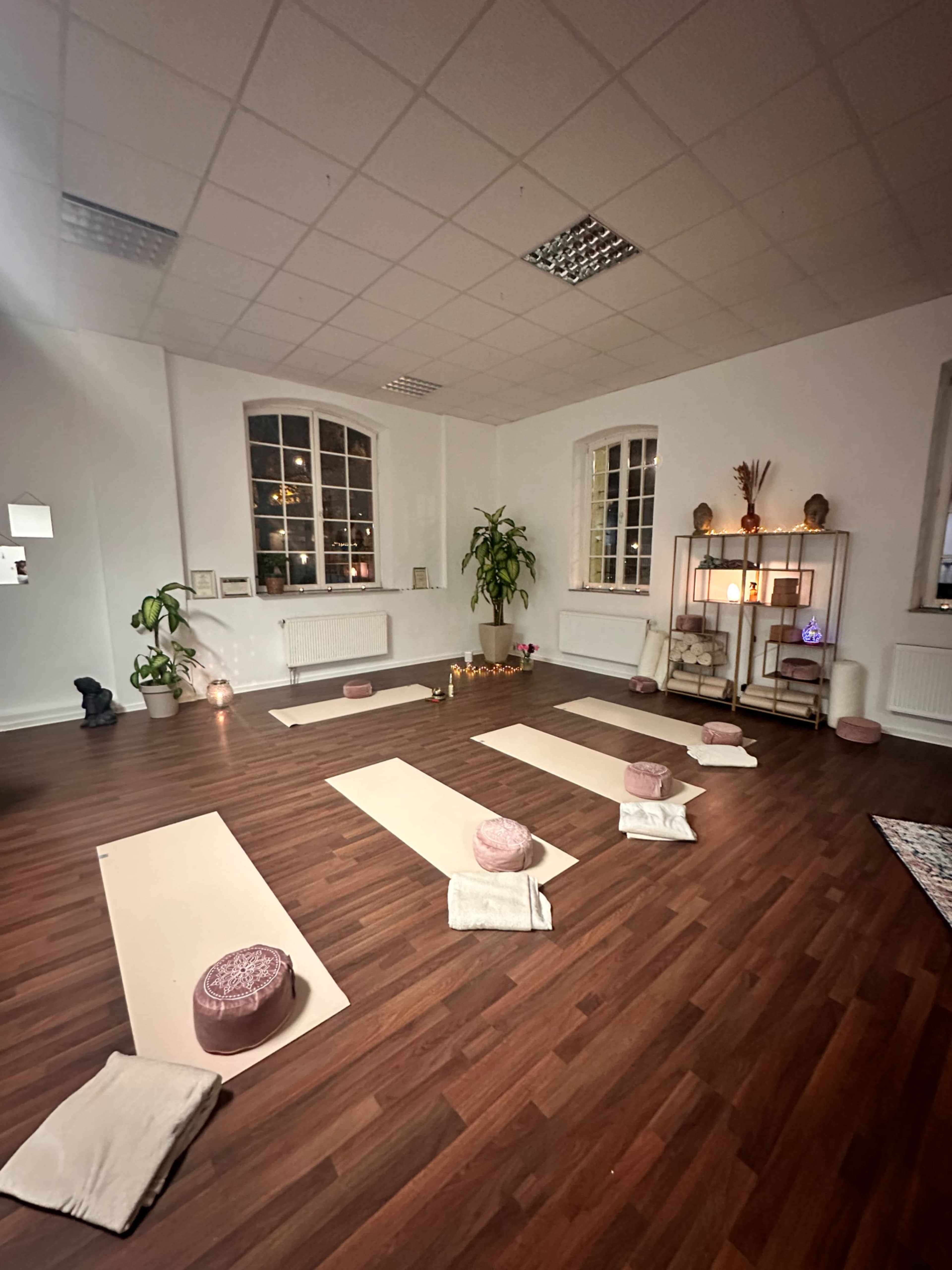 The image shows a yoga studio with several yoga mats arranged on the floor, surrounded by decor including cushions, plants, and a shelf with candles and decorative items.