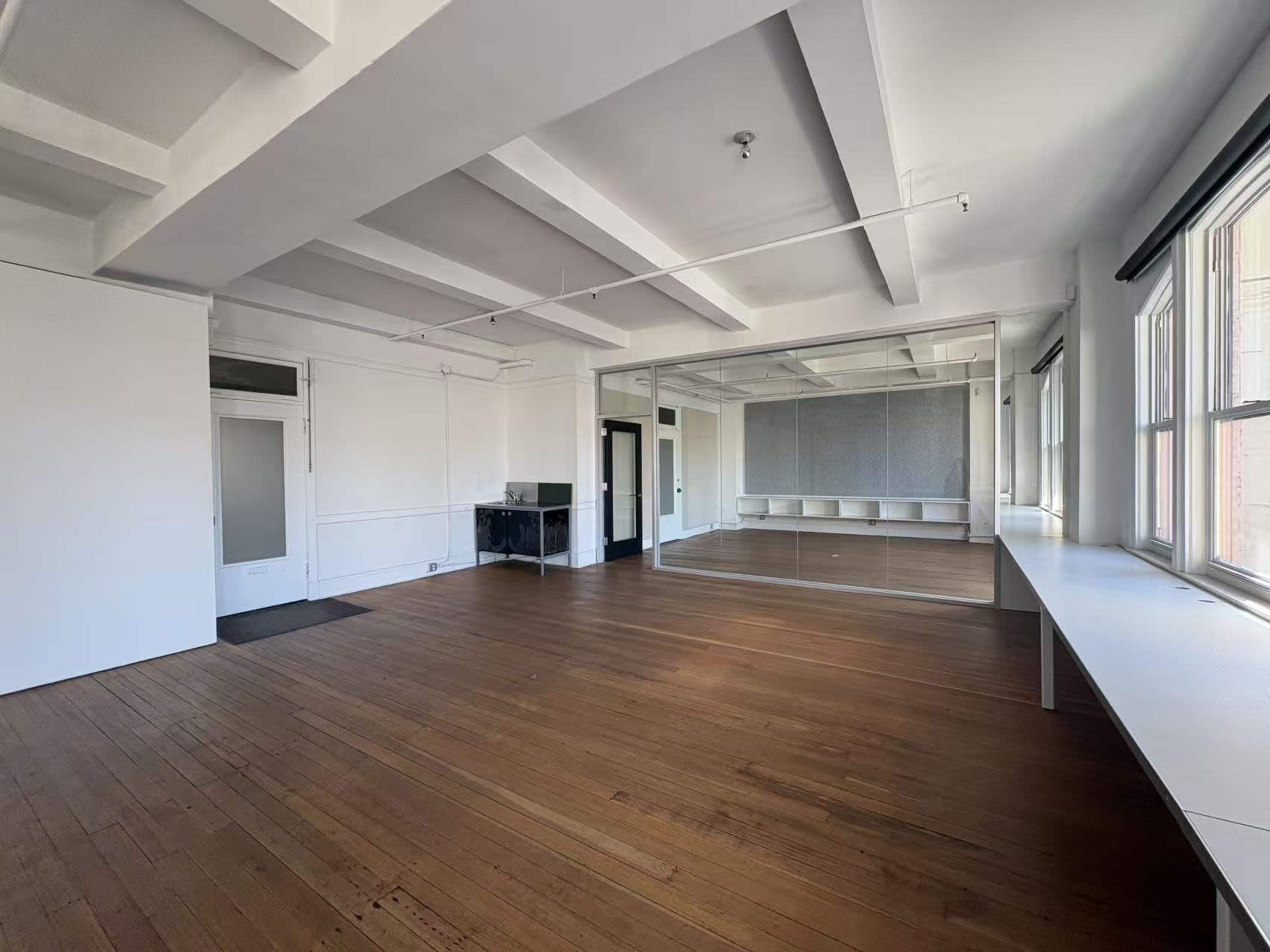 The image shows a spacious, uncluttered room with wooden floors, white walls, and large windows on one side.