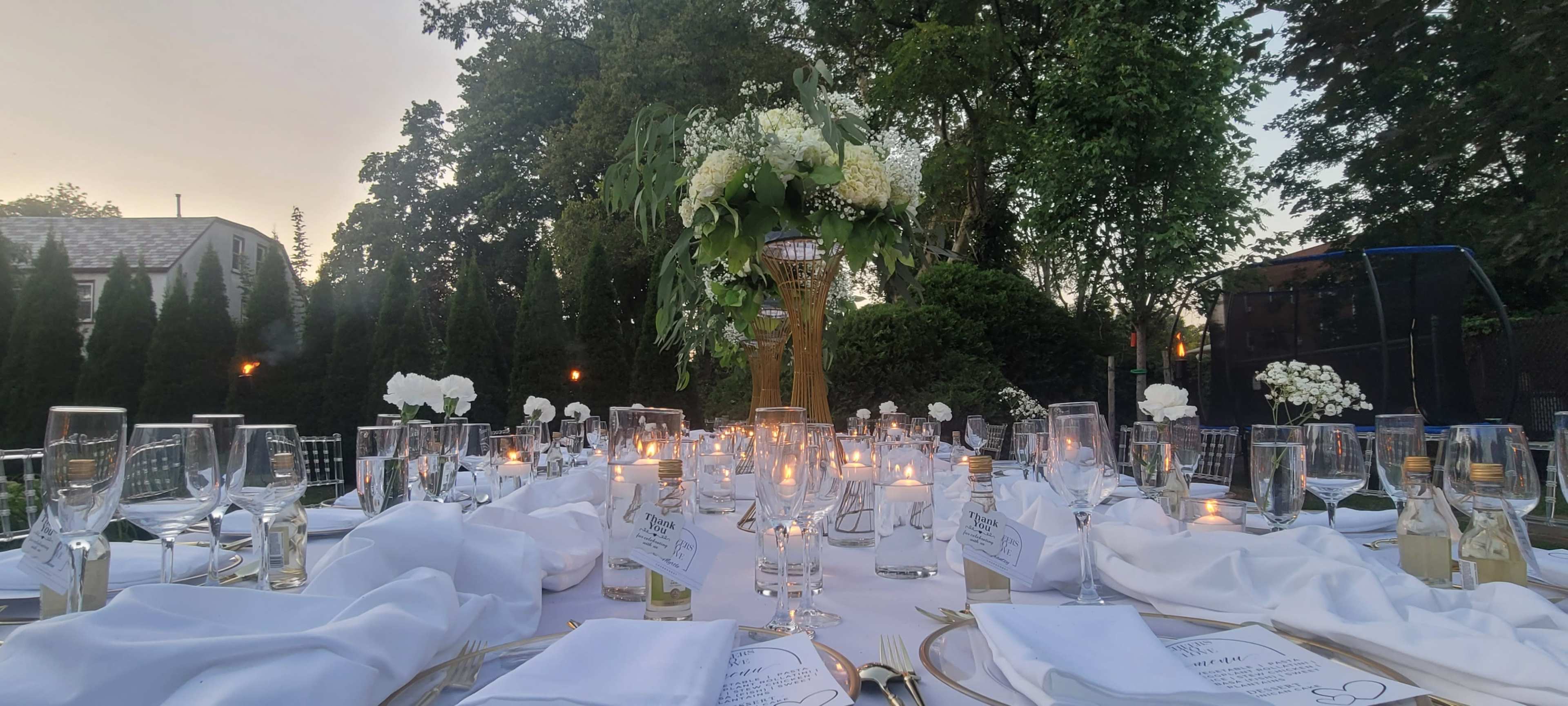 A beautifully arranged outdoor dining table is set with elegant white linens, glassware, and floral centerpieces, illuminated by soft candlelight.