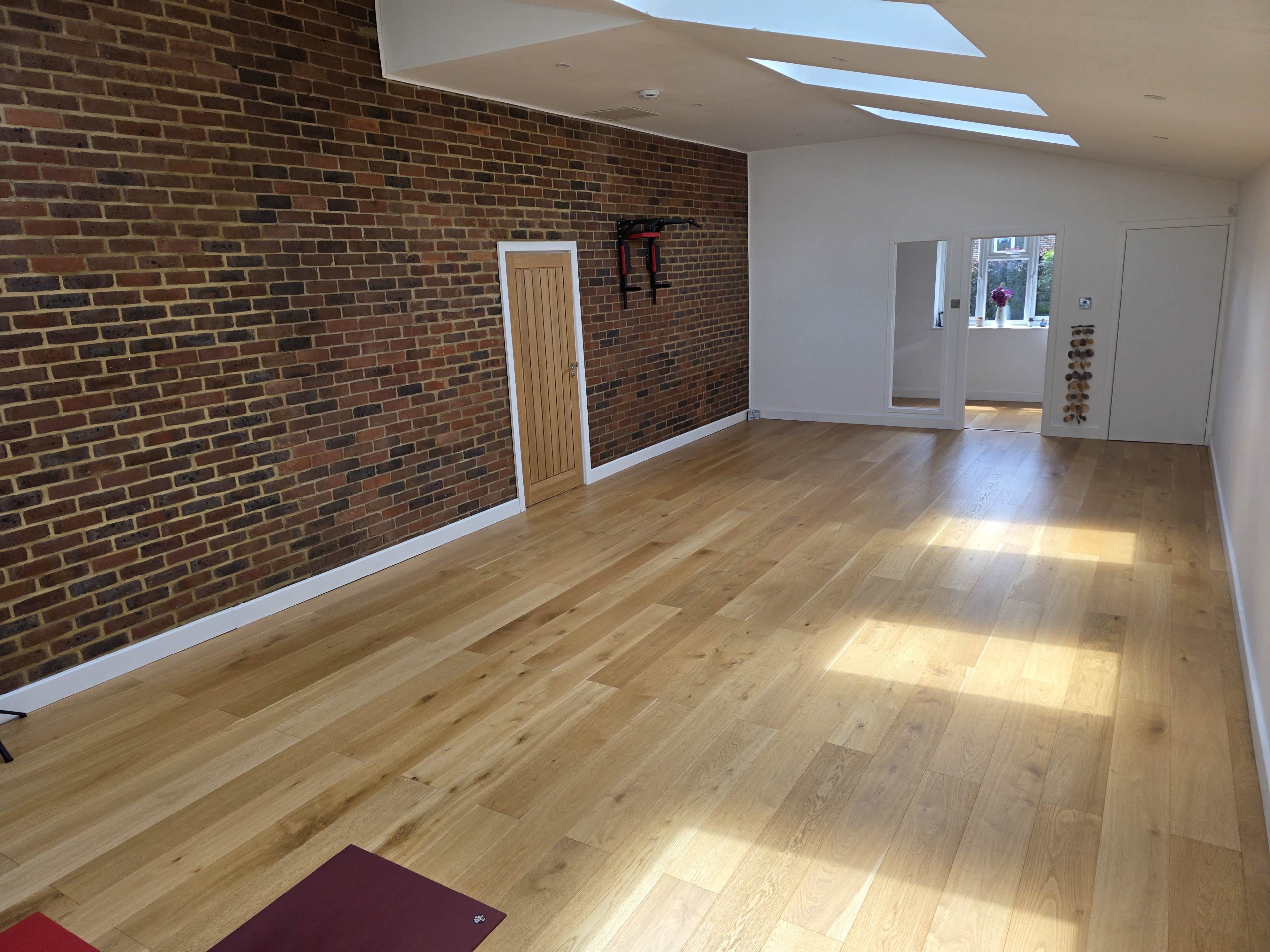 The image shows a spacious interior room with a wooden floor, brick walls, a door, mirrors, and a few exercise mats placed on the floor.