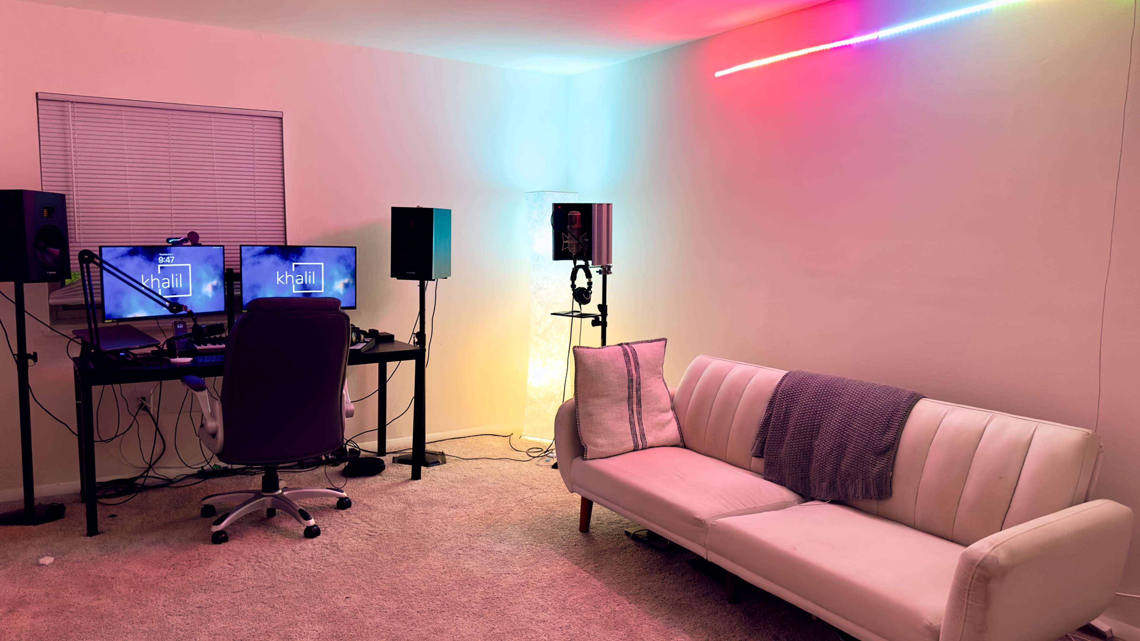 A modern workspace with two computer monitors on a desk, a microphone set up, and a white couch against a wall illuminated by colored lights.