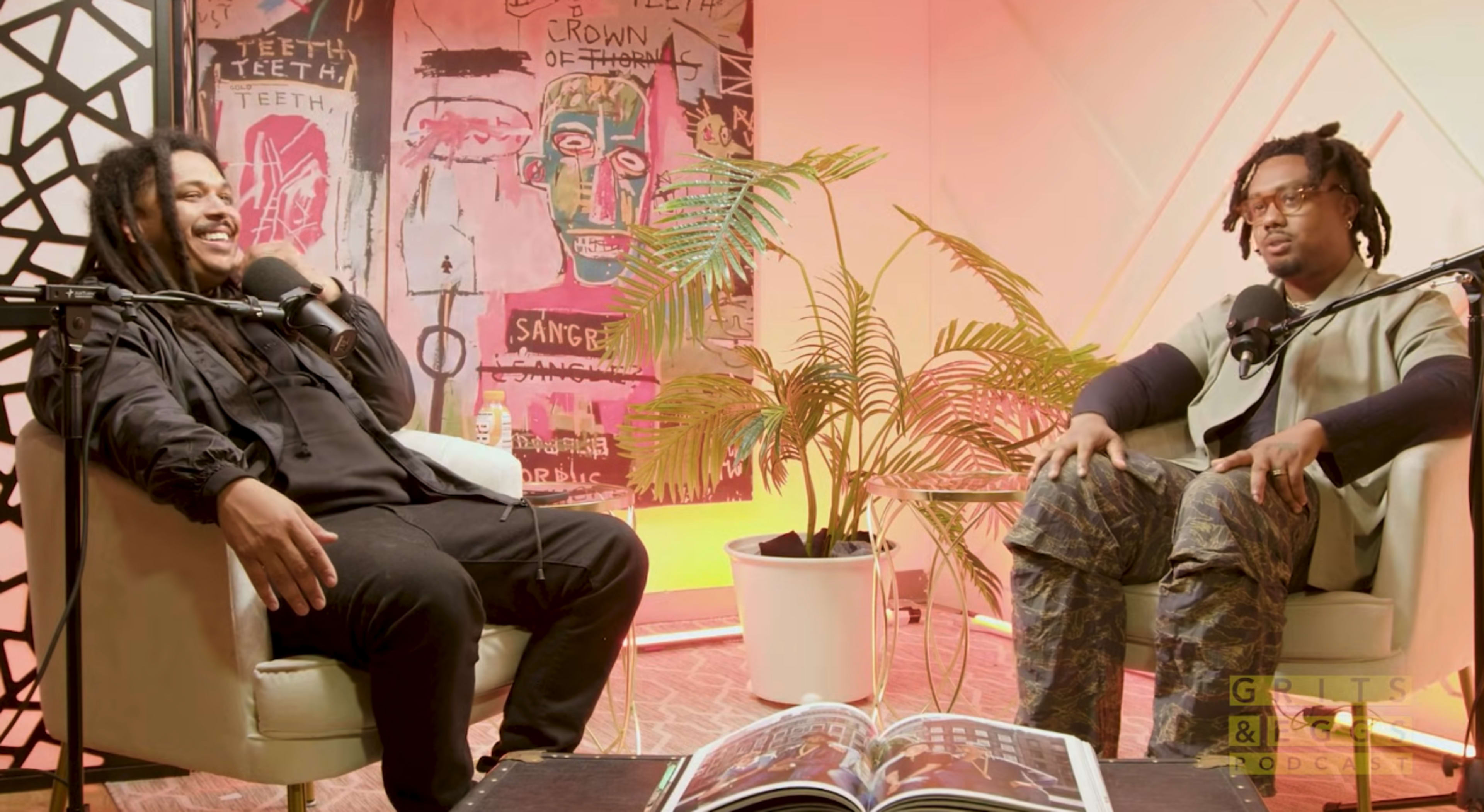 Two men sit in armchairs in a brightly lit room adorned with colorful artwork, engaged in conversation while a potted plant sits between them.