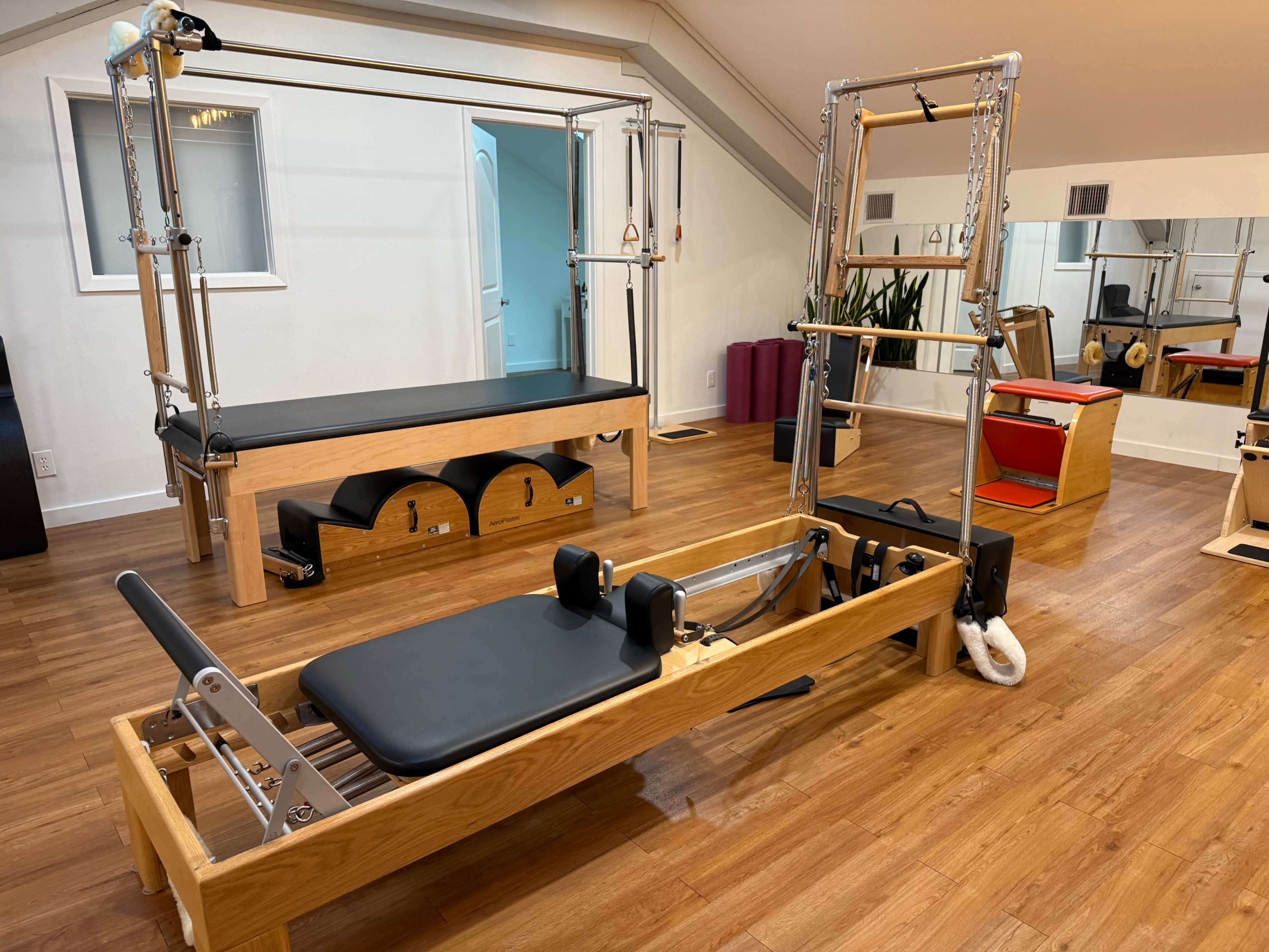 The image shows a Pilates studio with two equipment structures: a reformer in the foreground and a trapeze table in the background, both set on a wooden floor.