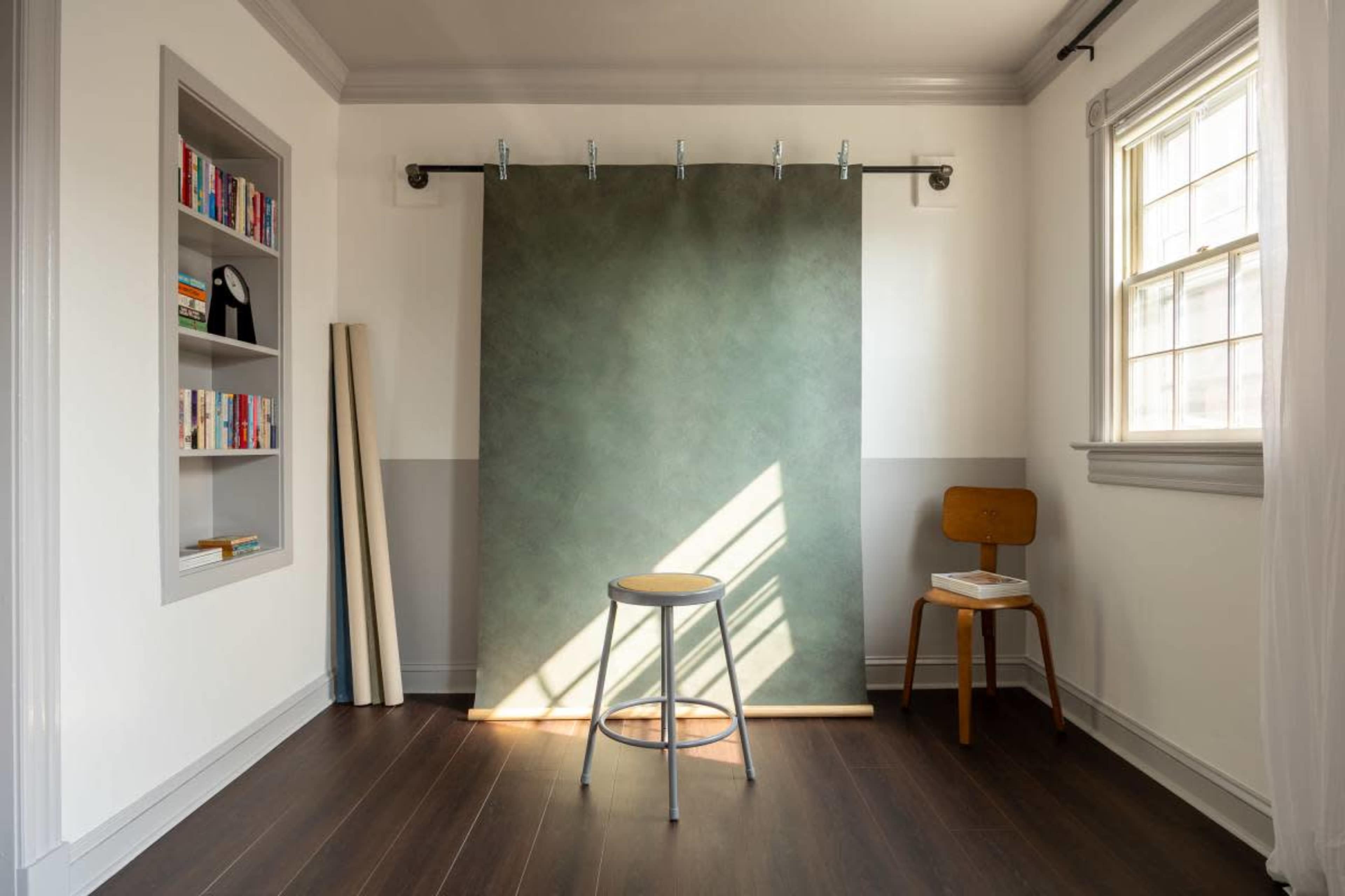 The image shows a well-lit room featuring a green backdrop on a track, a stool in front, and a bookshelf on the side with books.
