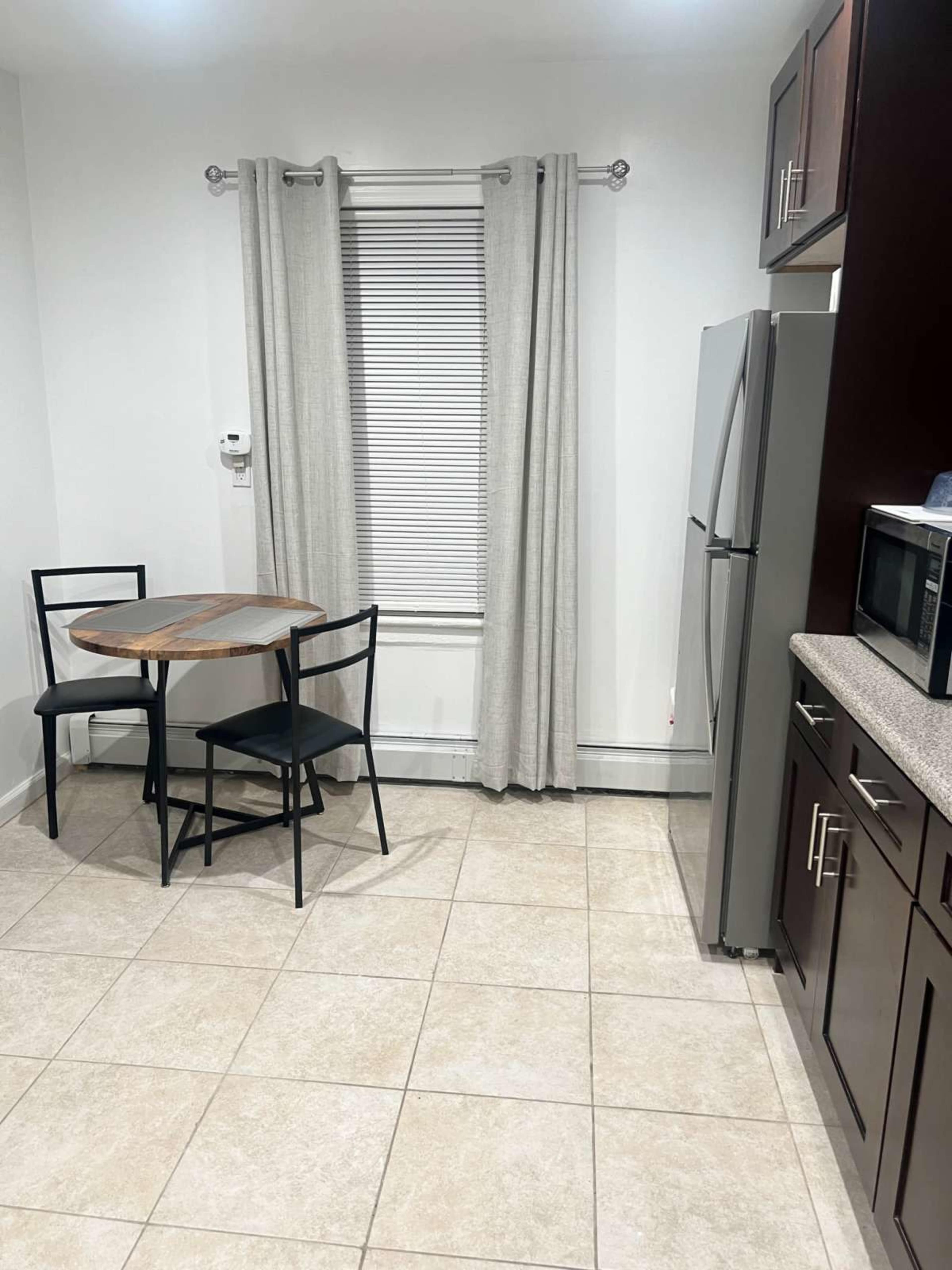 A kitchen featuring a small round table with two black chairs, a window covered with light curtains, a stainless steel refrigerator, and a microwave on the counter.
