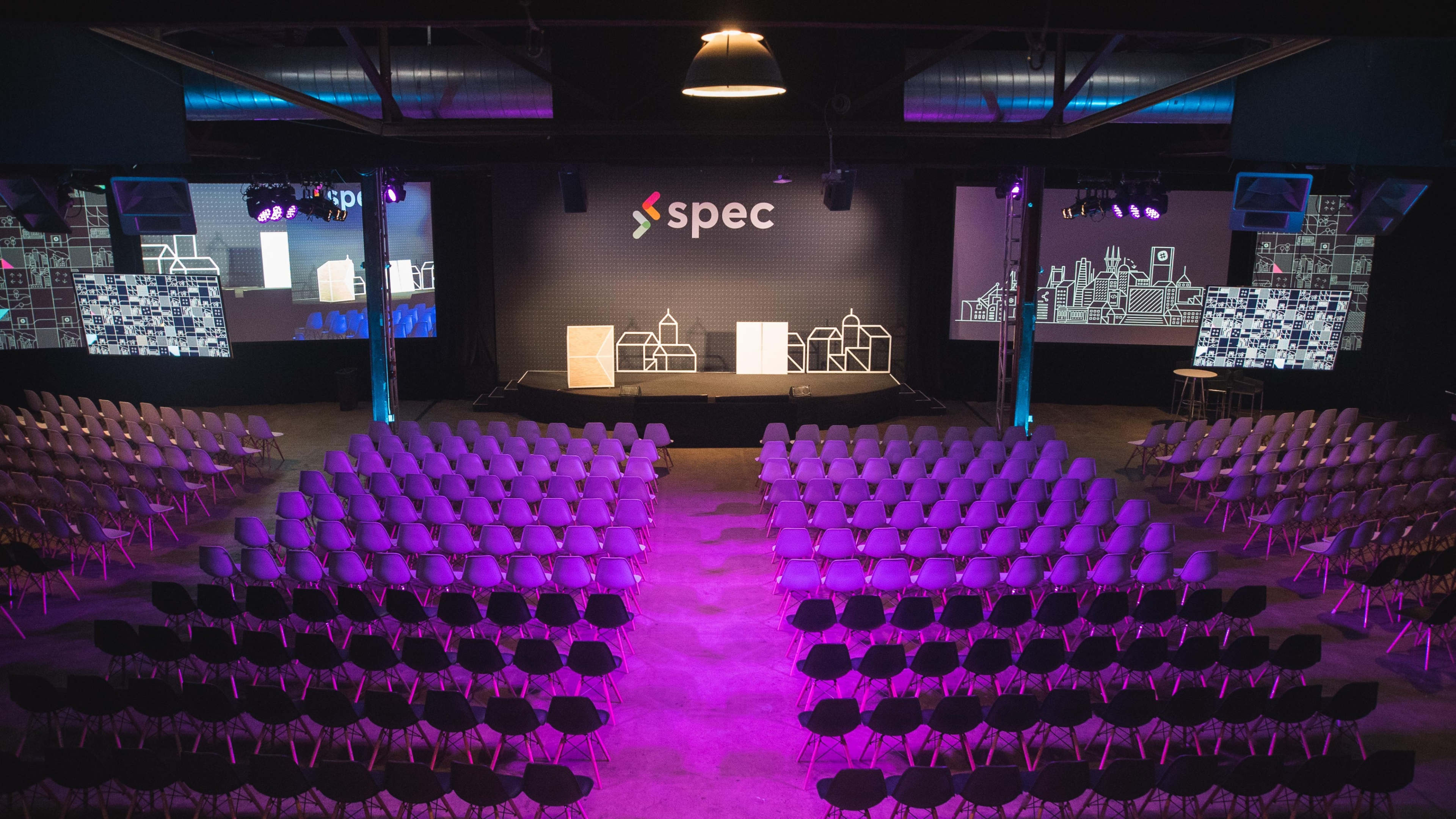The image shows an empty auditorium set up for an event, featuring rows of chairs facing a stage with a backdrop that includes the word "spec" and large screens displaying digital graphics.