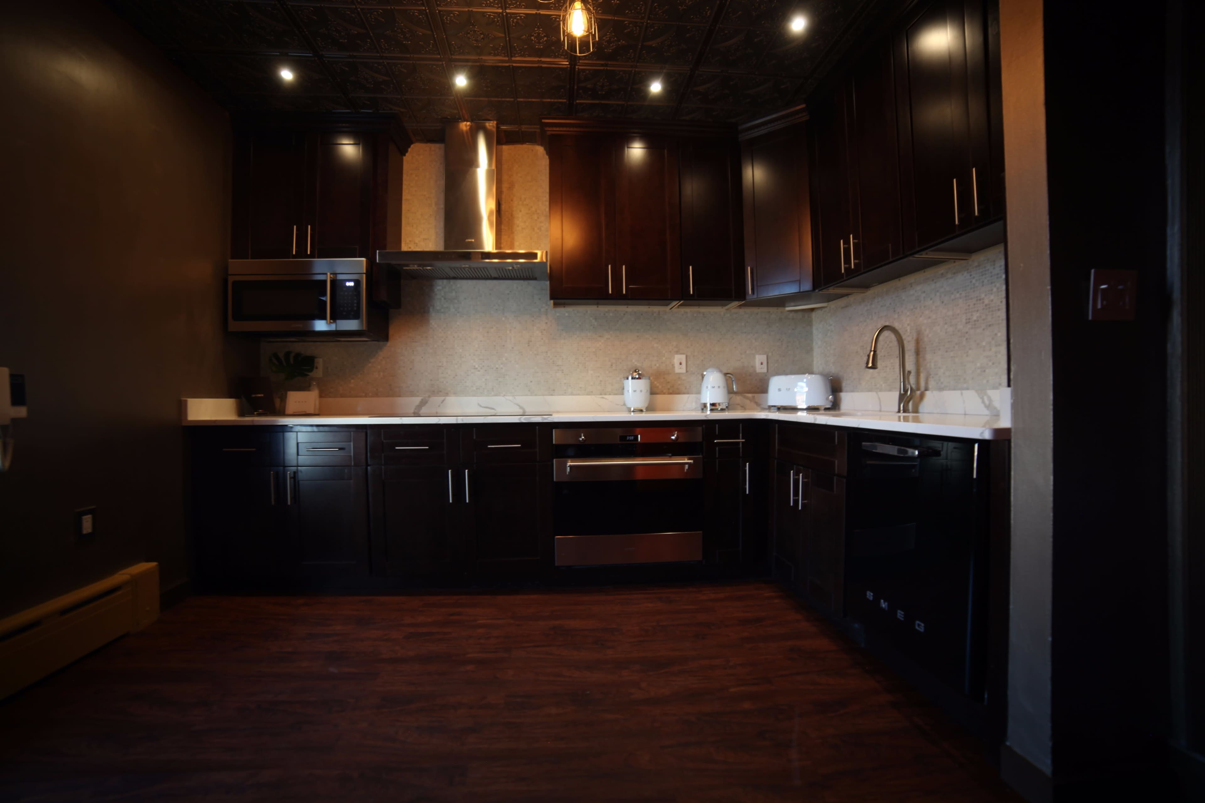 The image shows a modern kitchen with dark wooden cabinets, stainless steel appliances, and a white countertop illuminated by overhead lights.