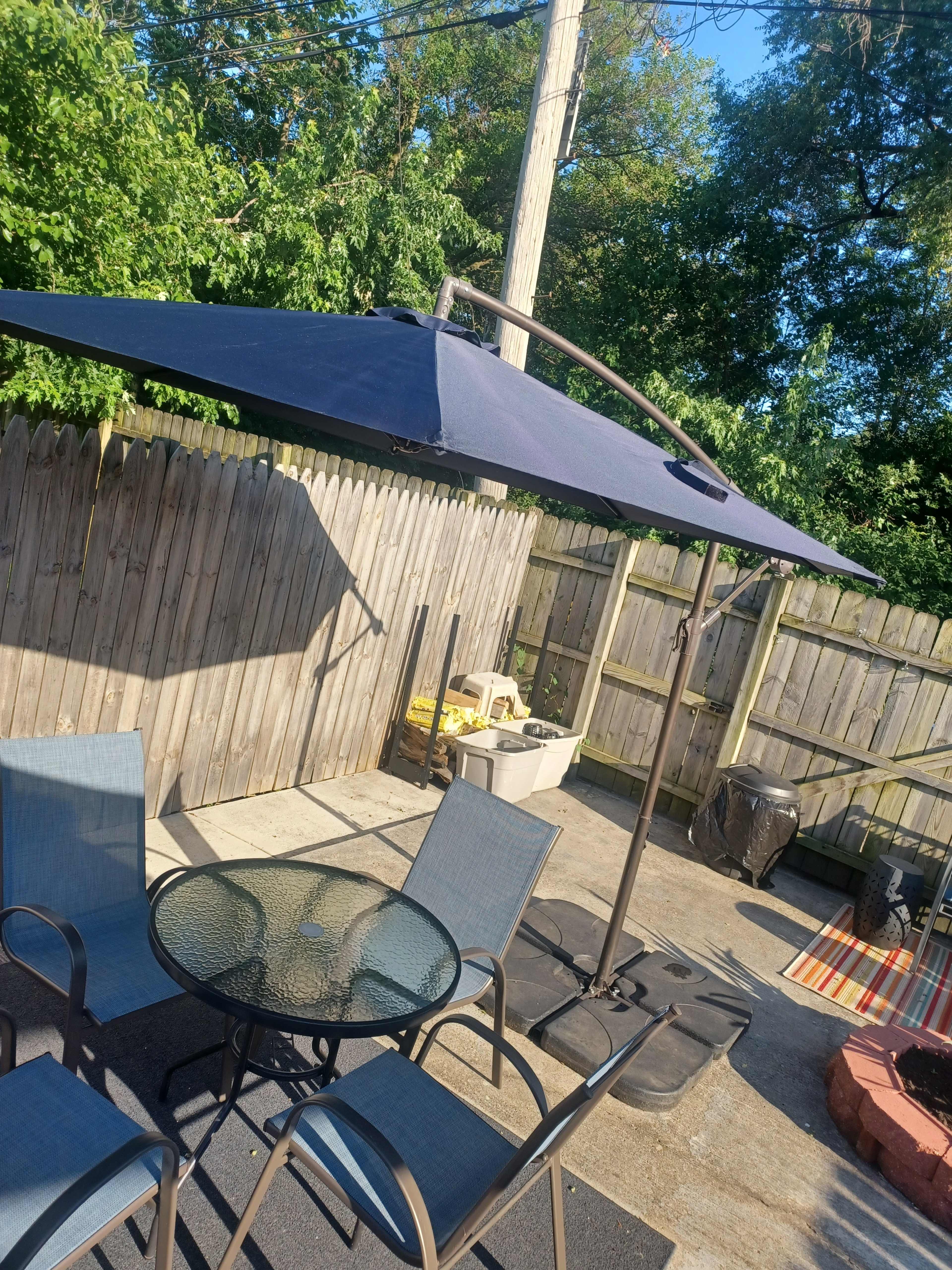 A patio area features a round glass table surrounded by four chairs, with a large umbrella positioned overhead and a wooden fence in the background.