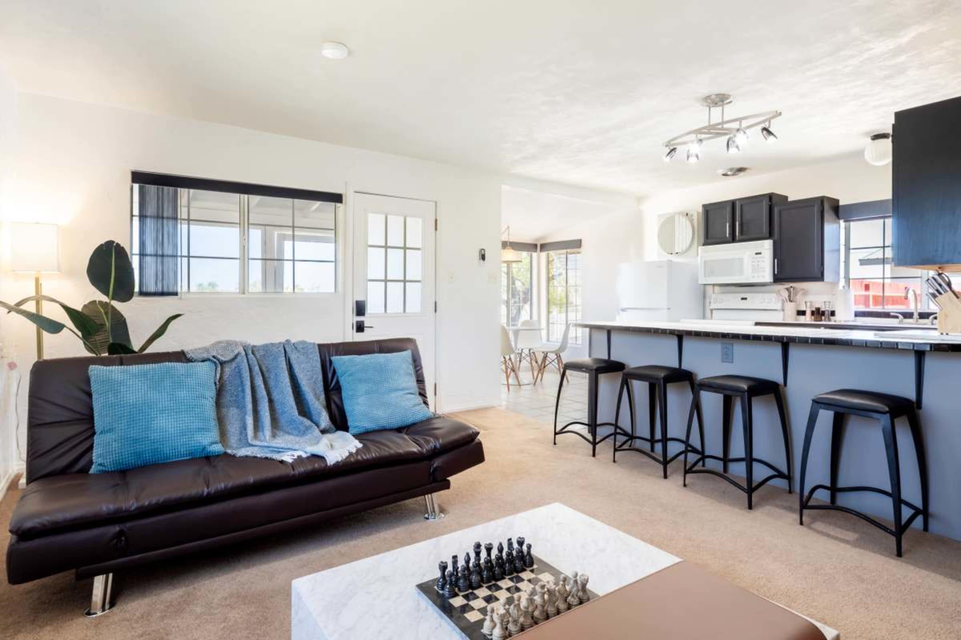 The image shows a modern, spacious living area featuring a sofa, a coffee table with a chess set, and a kitchen bar with stools in the background.
