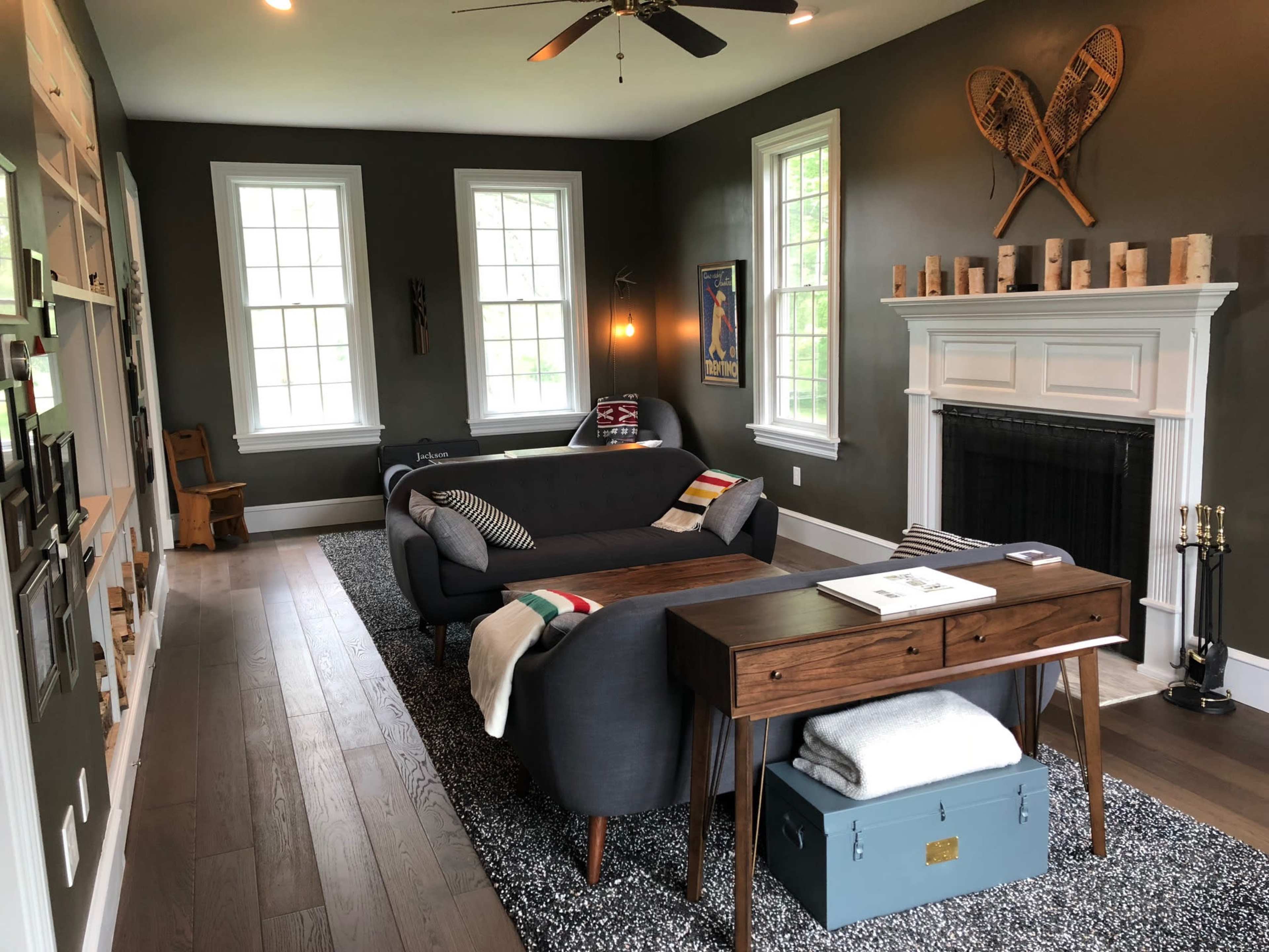 A modern living room featuring two gray sofas, a wooden table, a fireplace, and decorative wall art in a space with dark green walls and large windows.
