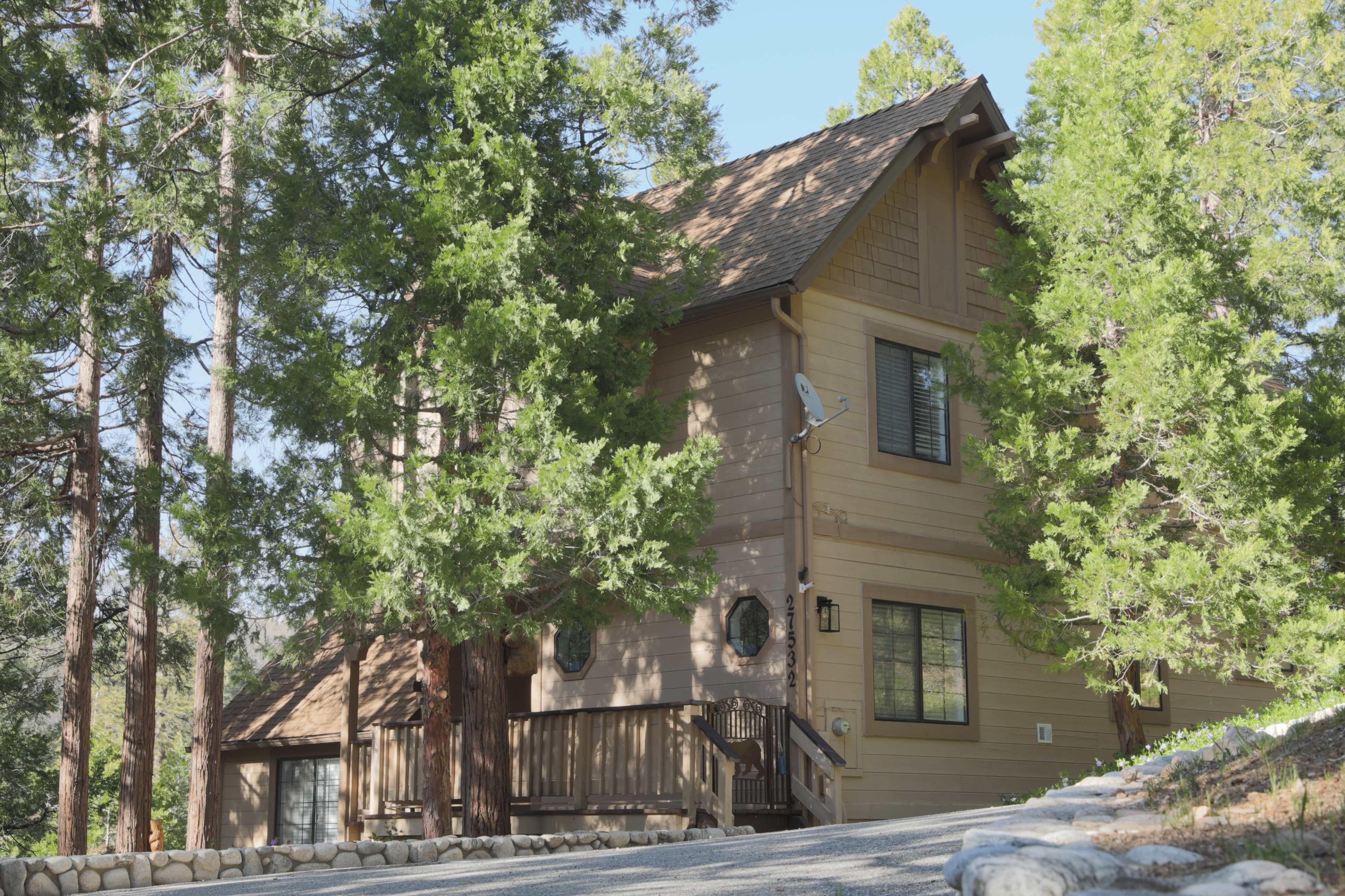 A two-story wooden cabin is situated among tall trees on a gravel road.