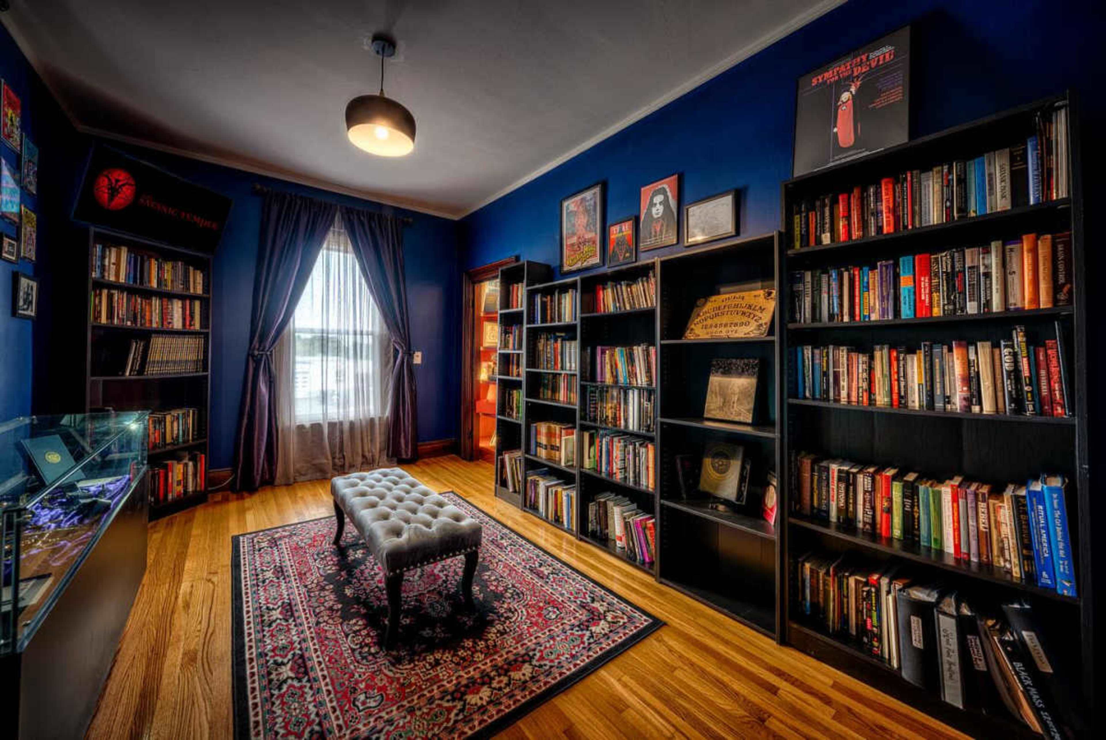 The image shows a cozy library room featuring dark blue walls, tall bookshelves filled with various books, a tufted bench in the center, and a window with drapes allowing natural light to enter.