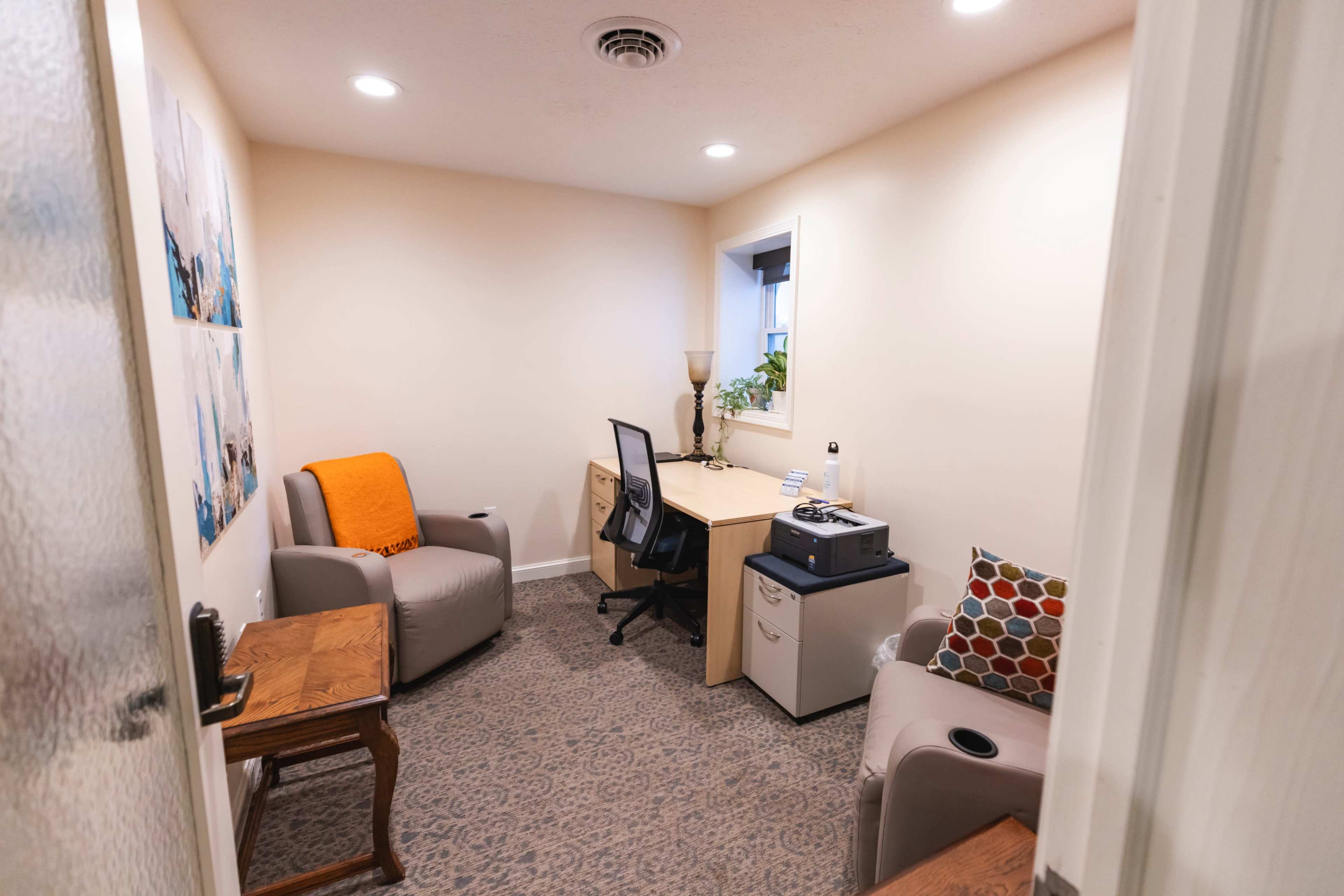 A small office features a desk, two chairs, and decorative items along with a doorframe visible in the foreground.