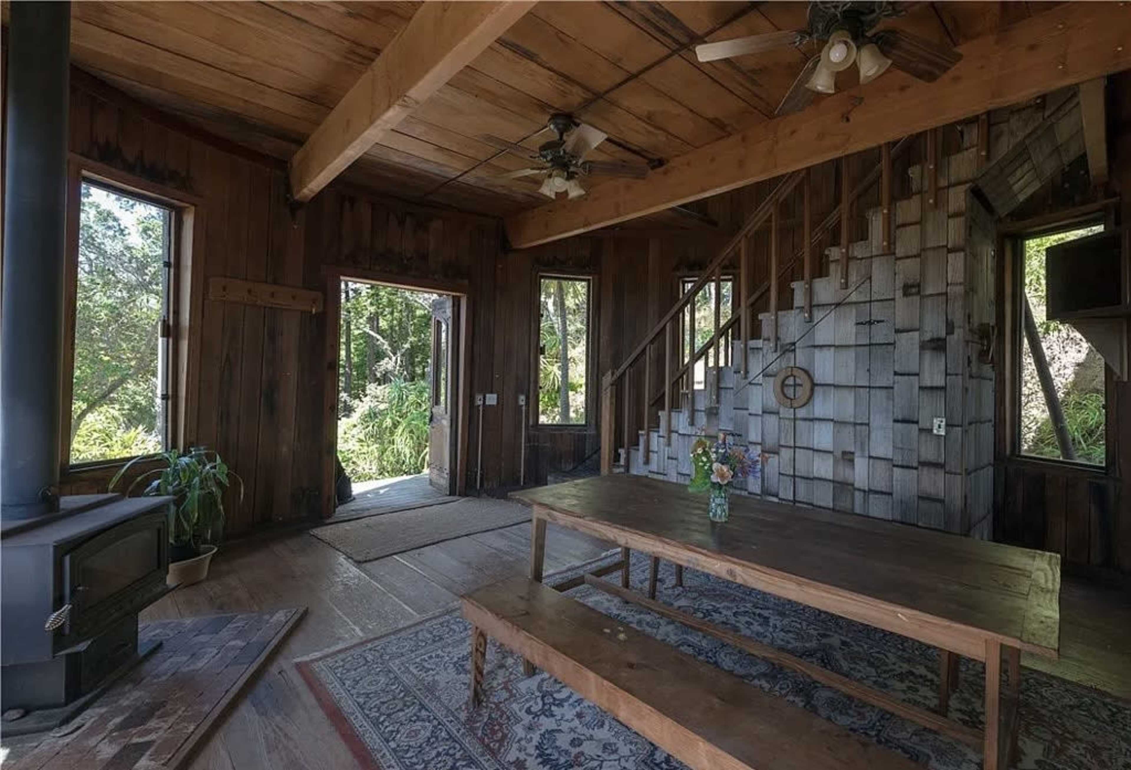 The image shows a cozy wooden interior featuring a large table with a vase of flowers, a stove, and a staircase leading to a second level.