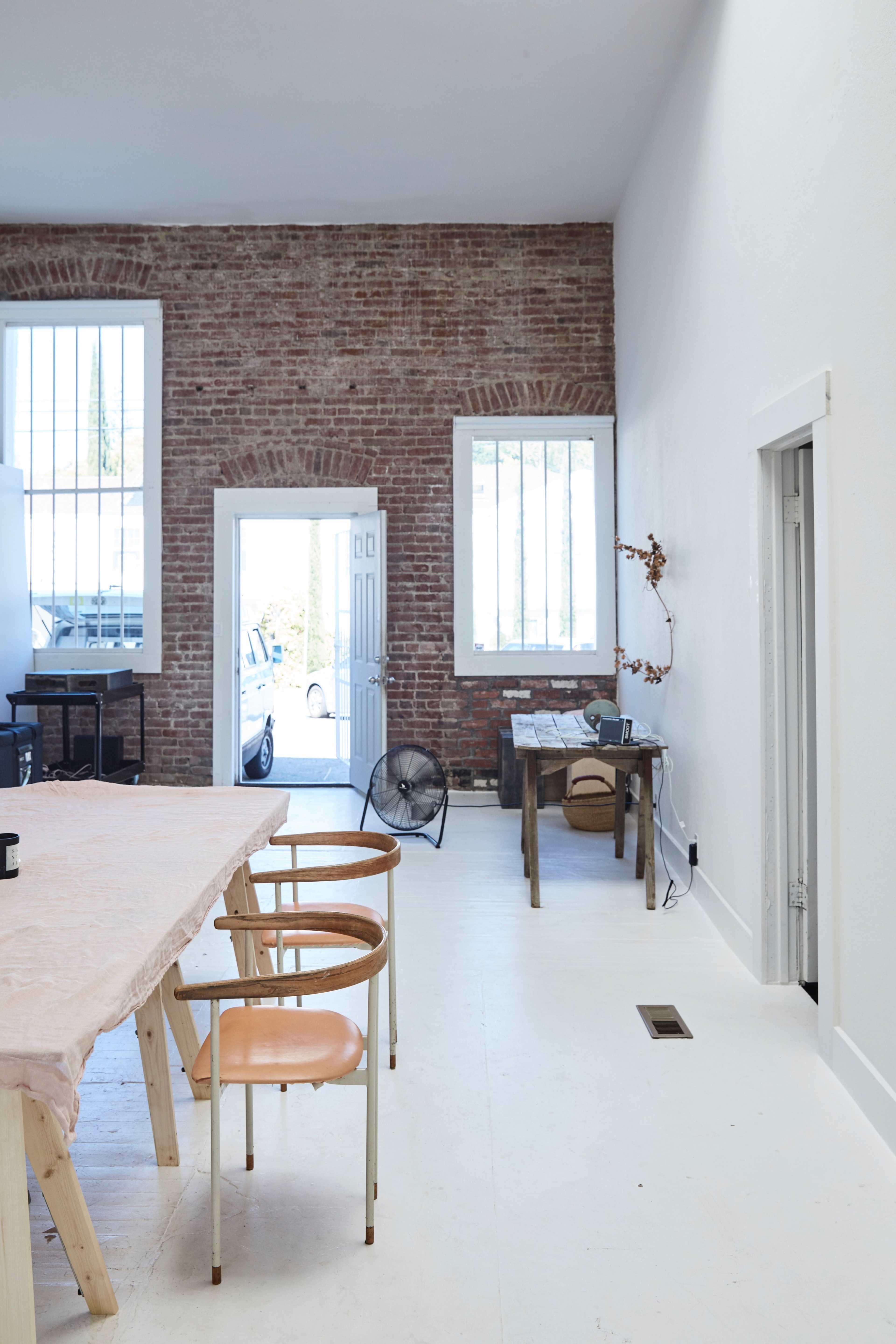 The scene features a spacious room with exposed brick walls, a long table with chairs, and an open door leading to an outdoor area.