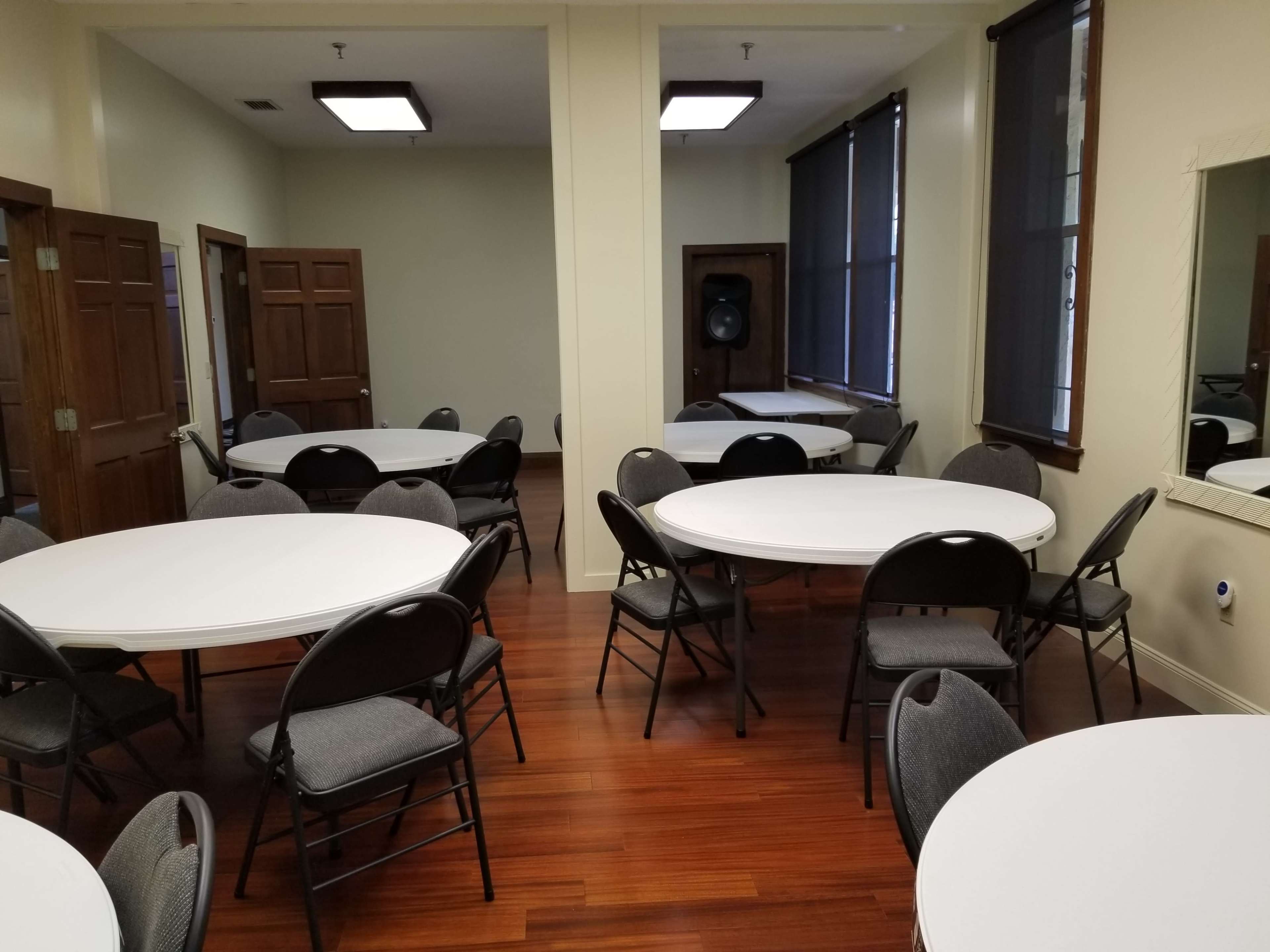 A room with several circular tables surrounded by chairs, featuring wooden floors and light-colored walls.