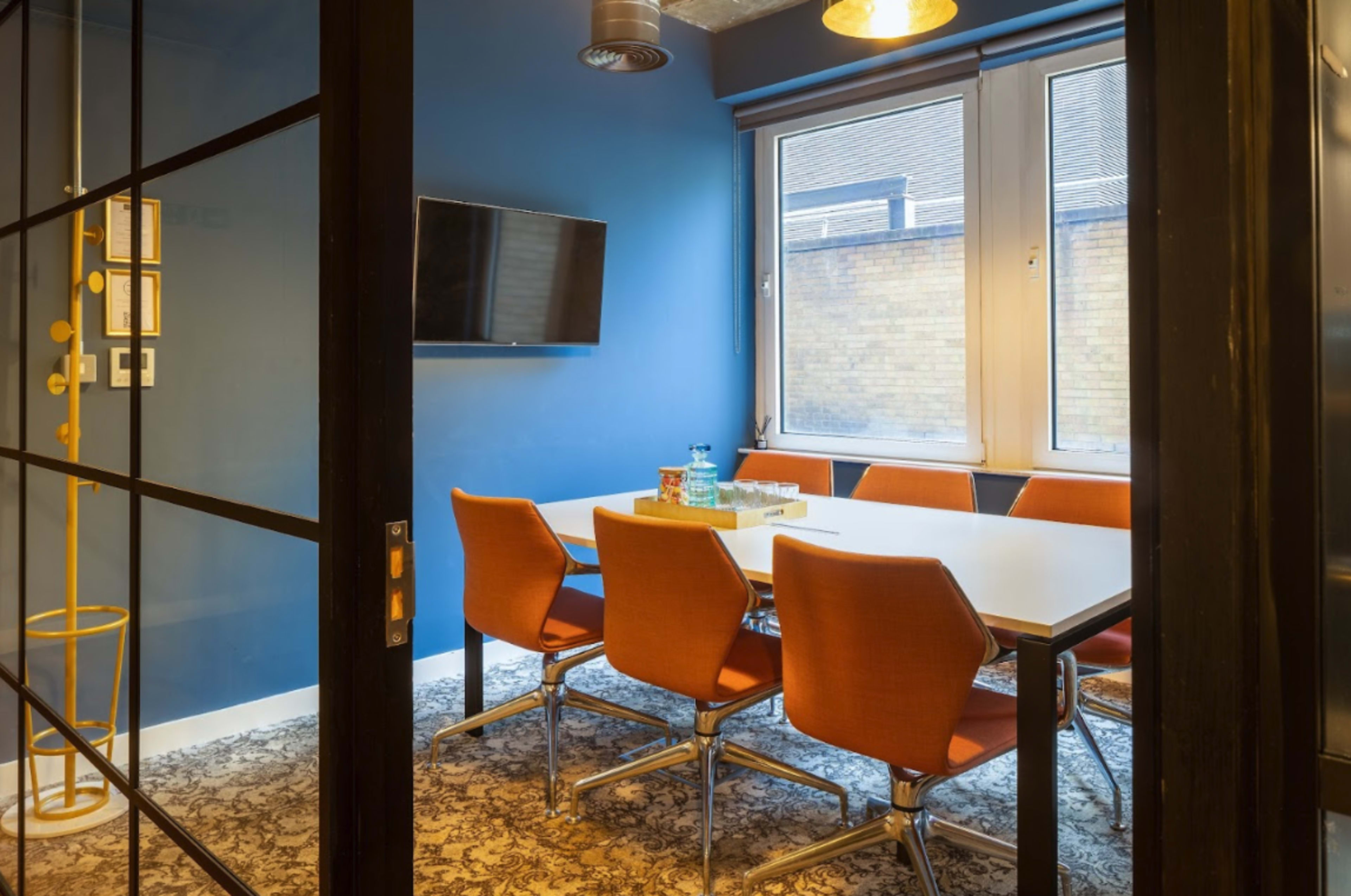 A meeting room features a large table surrounded by orange chairs, with a television mounted on the wall and large windows allowing natural light.
