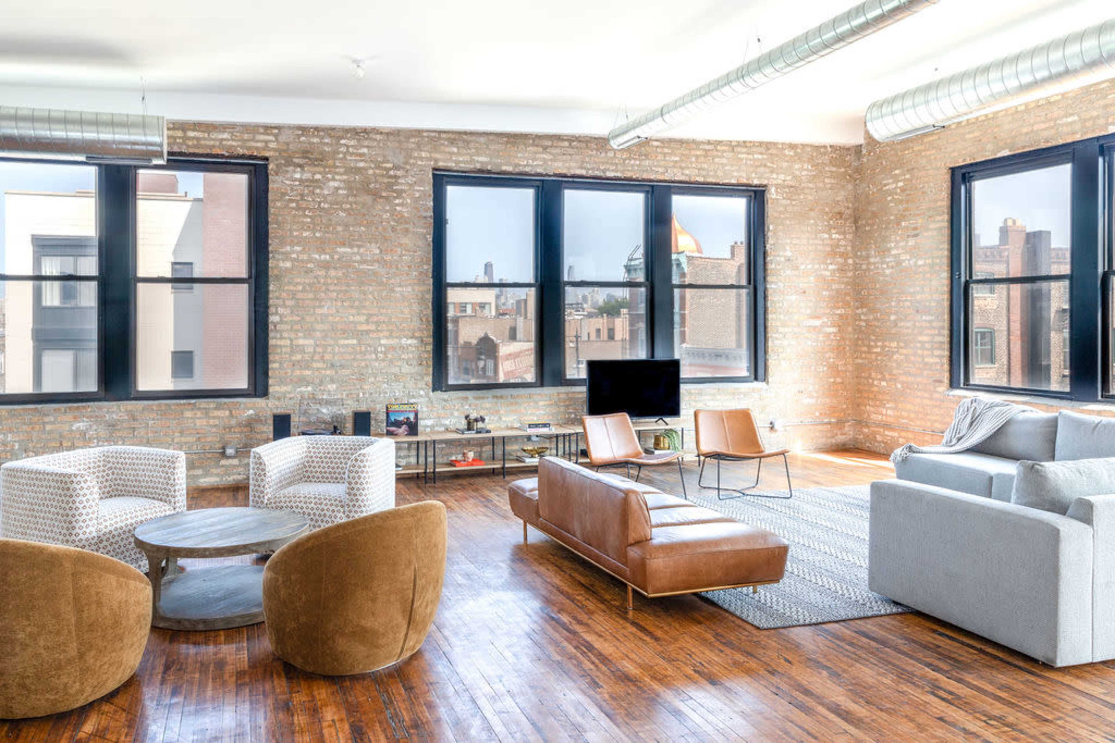 A spacious living room features large windows, exposed brick walls, and a mix of modern furniture including a sofa and armchairs.