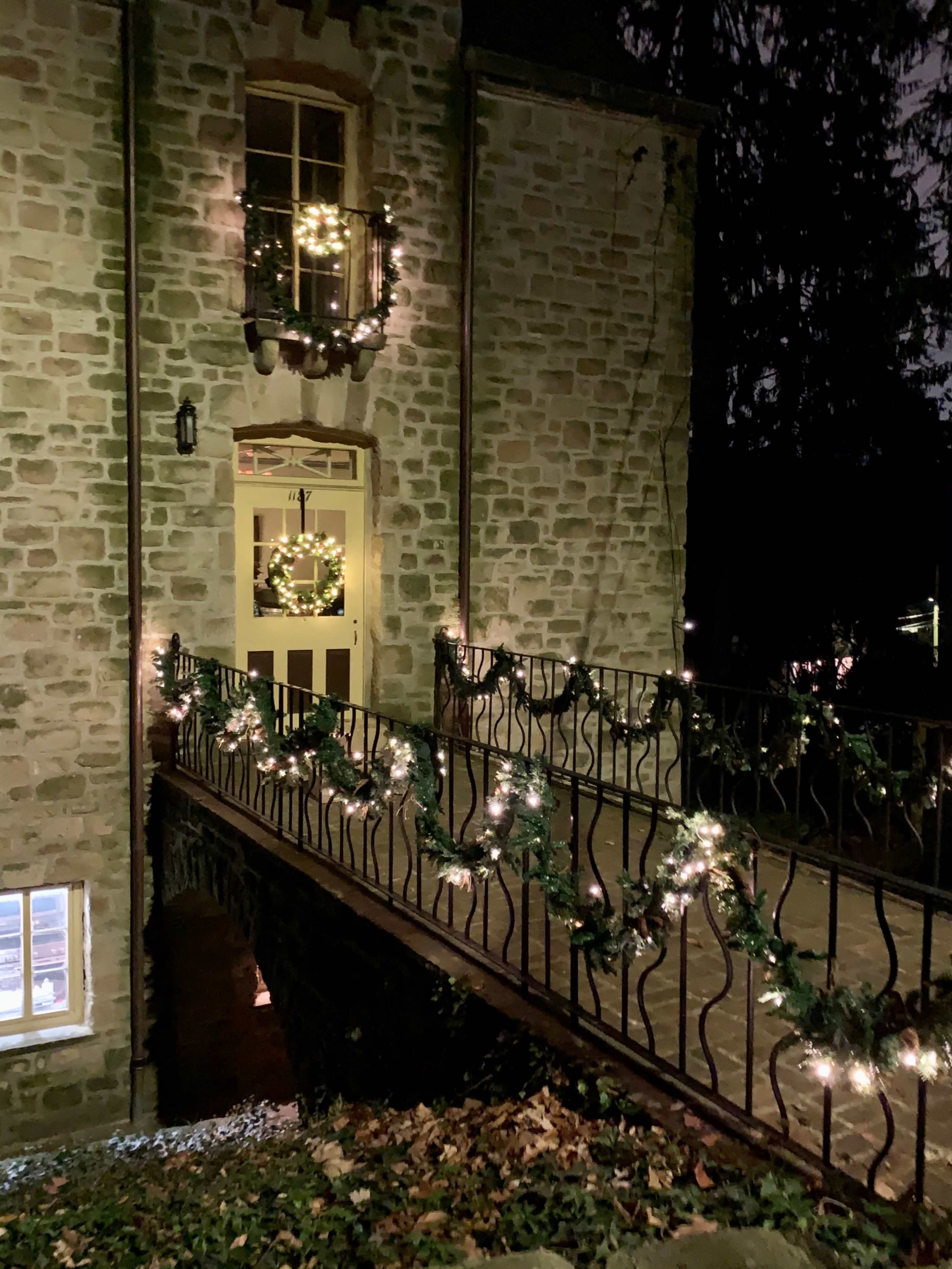 A stone building is illuminated at night with a decorated entrance and a railing adorned with garlands and lights.