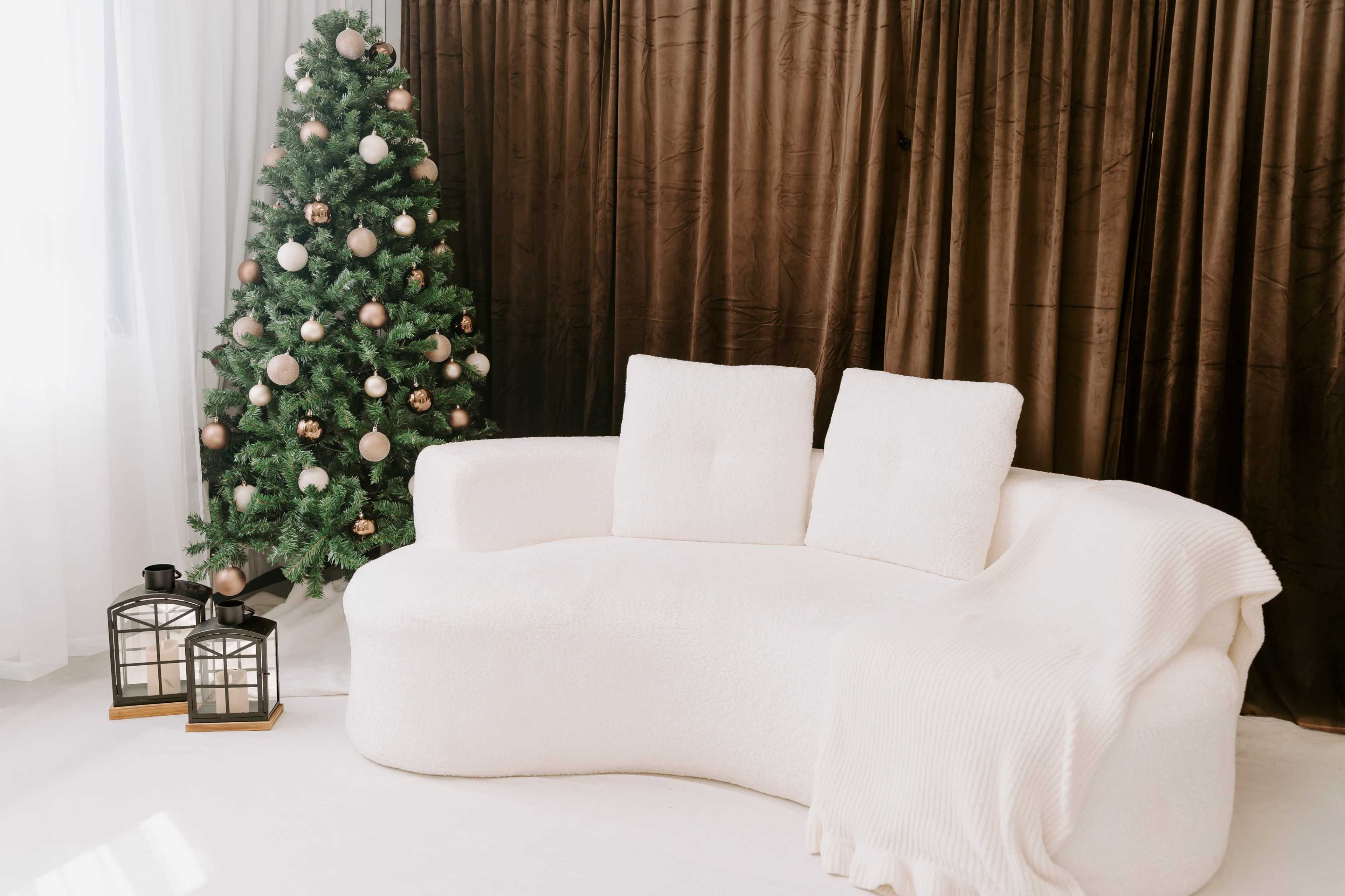 A white curved sofa with two cushions and a draped blanket is positioned beside a decorated Christmas tree in a softly lit room.