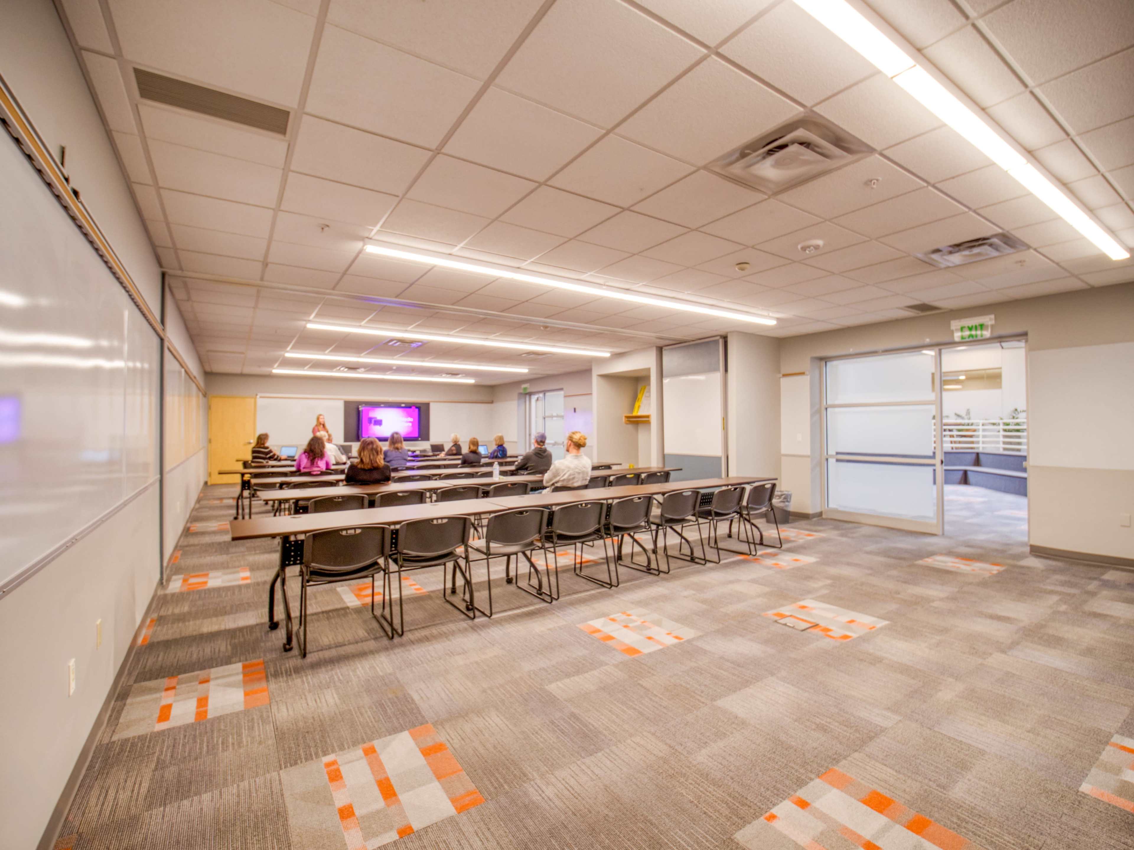 A spacious classroom features rows of chairs and a large screen at one end, with several people seated and facing the front.