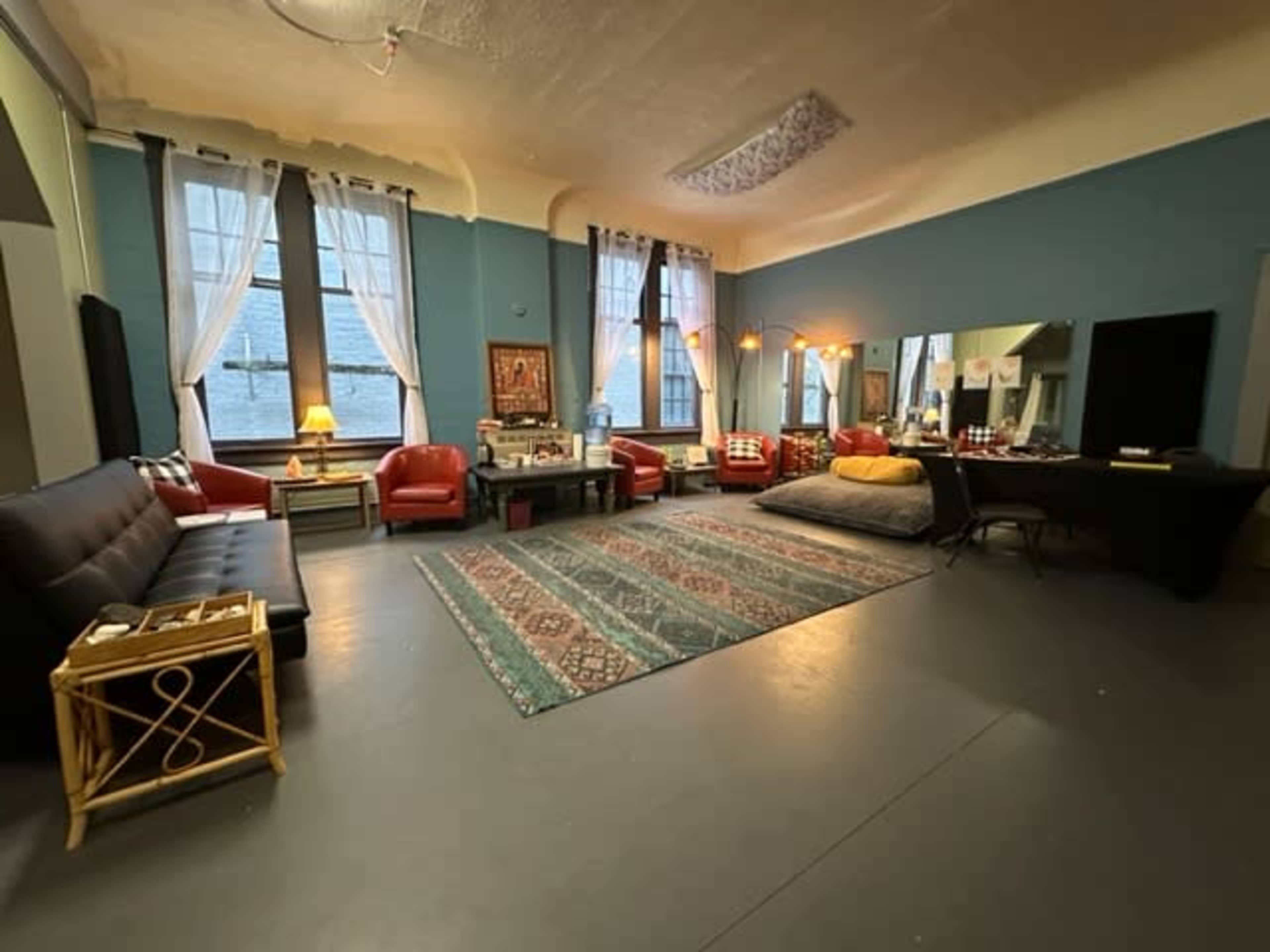 The image shows a spacious room with a large area rug, seating arrangements including a black sofa and red chairs, and large windows providing natural light.