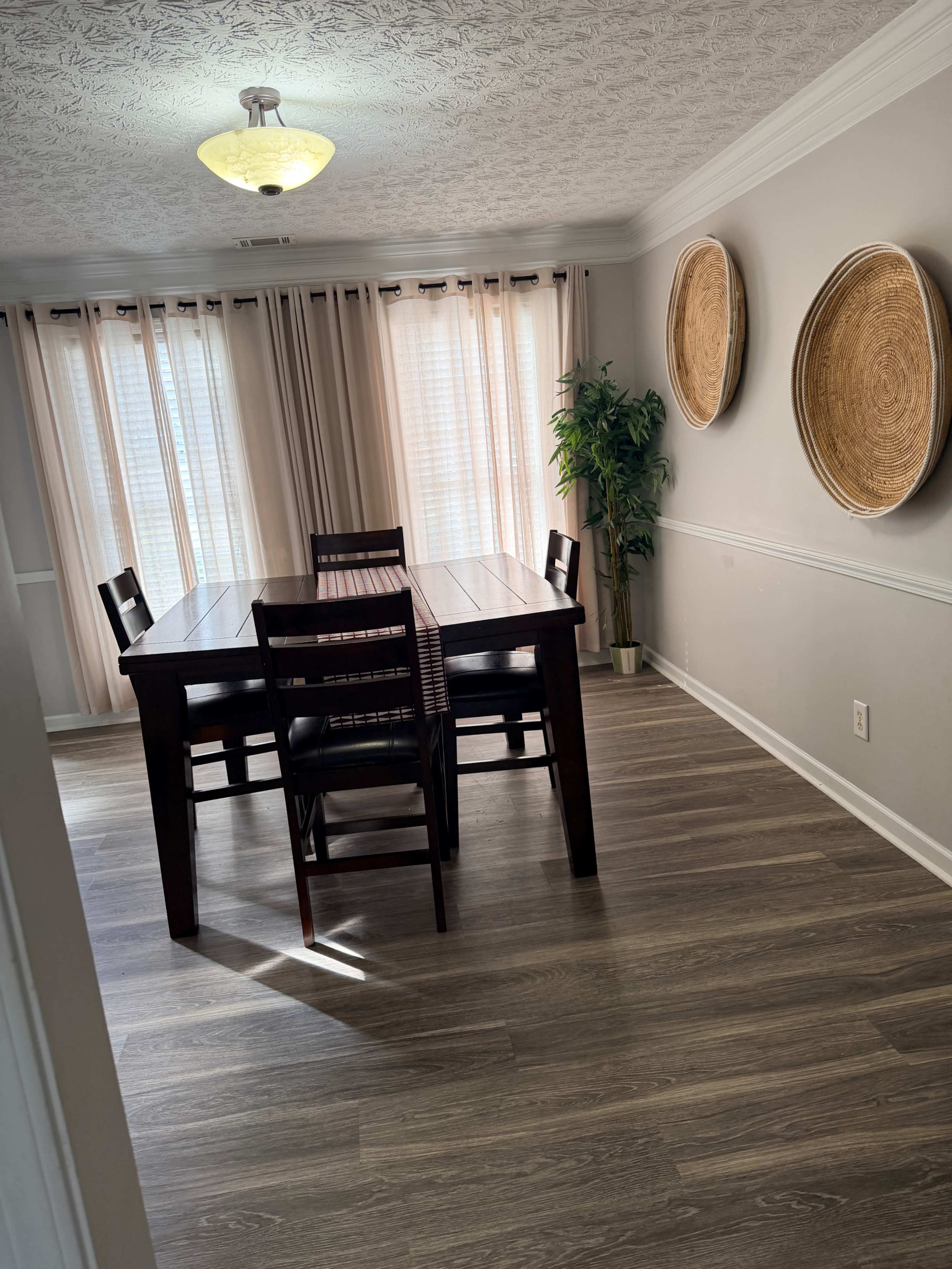 A dining room features a wooden table surrounded by four chairs, with natural light streaming through sheer curtains and two woven wall hangings.