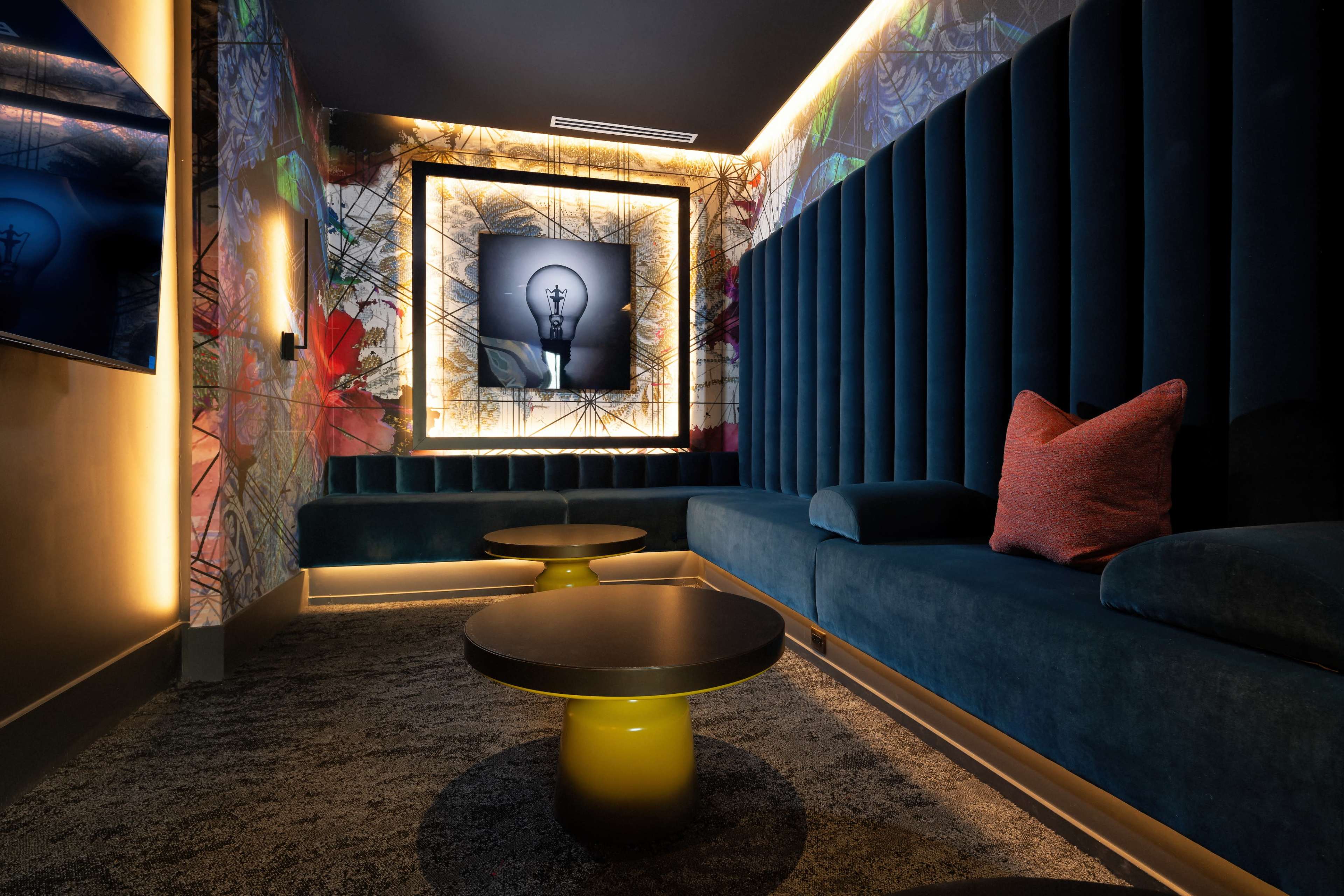 A modern interior lounge with plush blue seating, round black tables, and a bright lightbulb artwork framed on the wall.