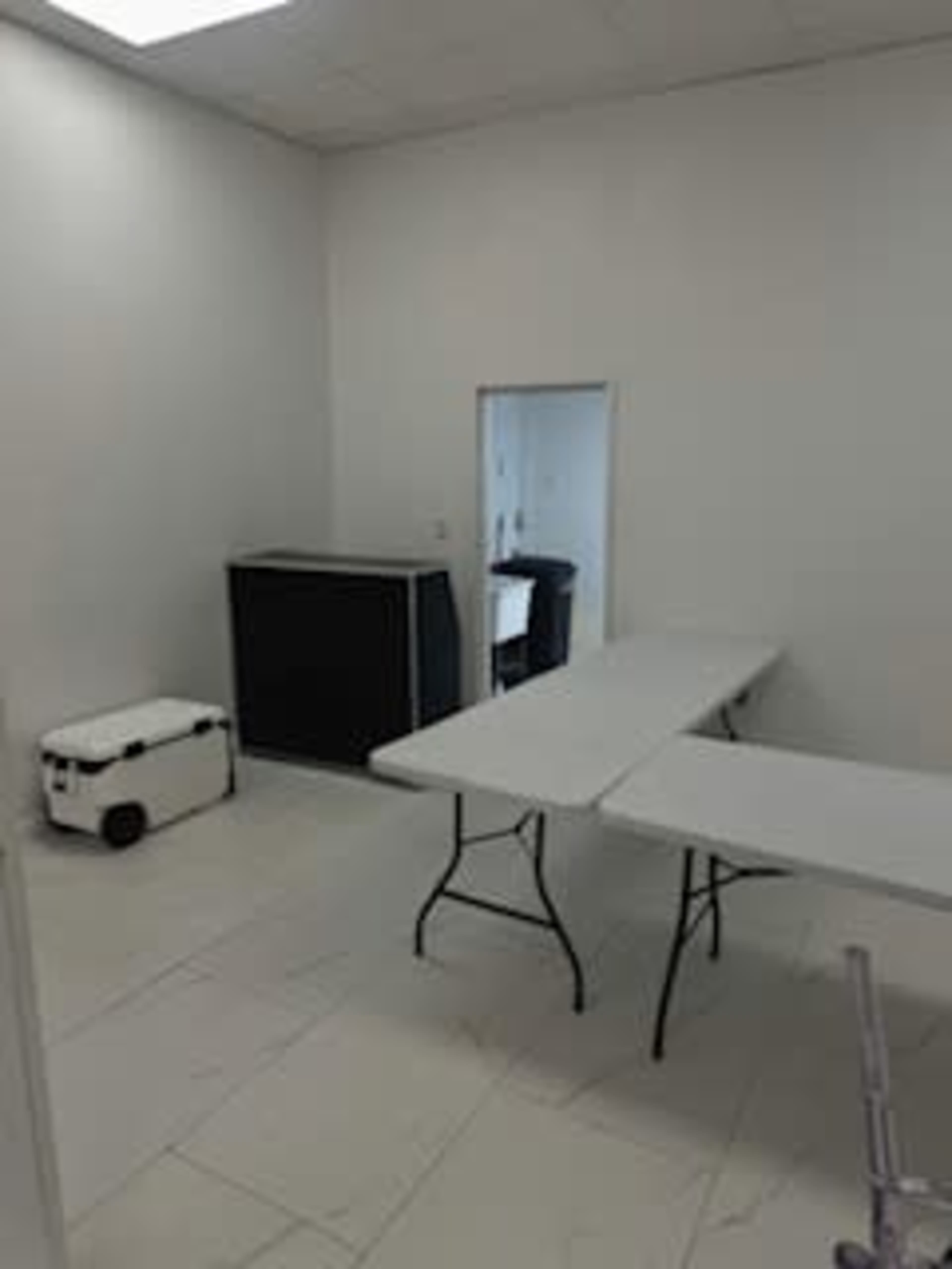 The image shows a sparsely furnished room with two folding tables, a cooler, and a black cabinet against a white wall.