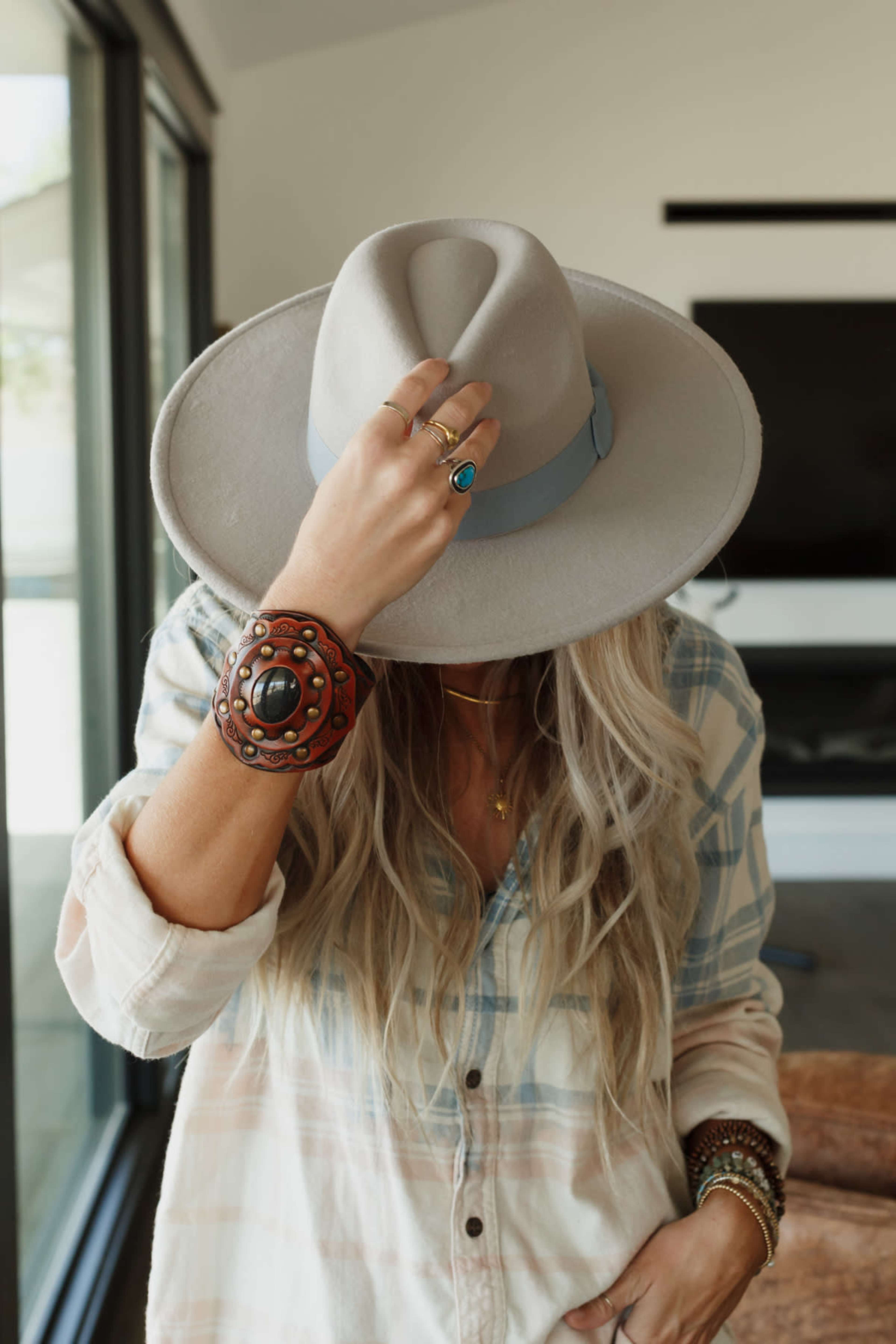 A person wearing a wide-brimmed gray hat and a patterned shirt stands indoors, with their head slightly bowed and their hand raised.