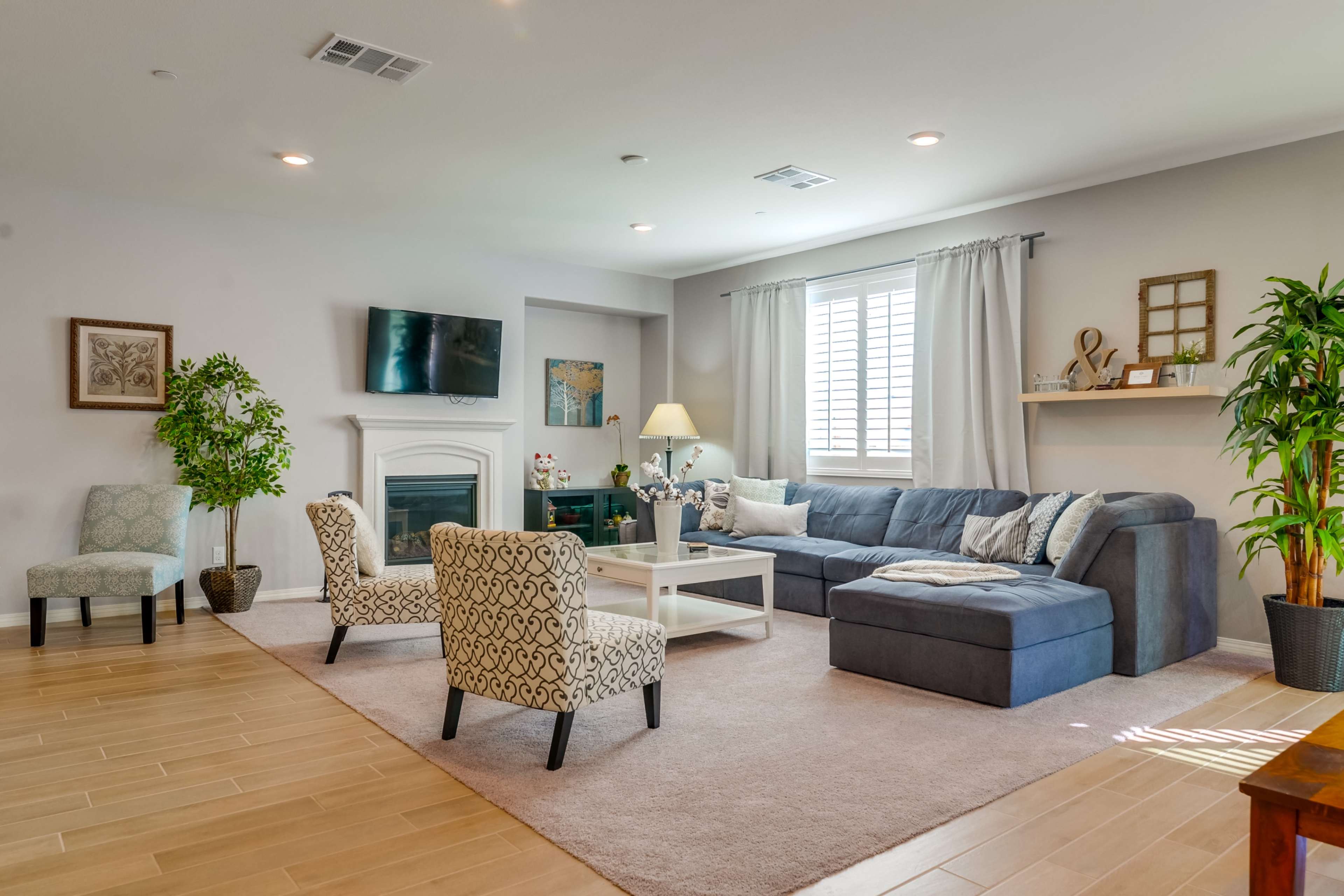A spacious living room with a blue sectional sofa, a coffee table, and two patterned chairs, along with a fireplace and a large window.