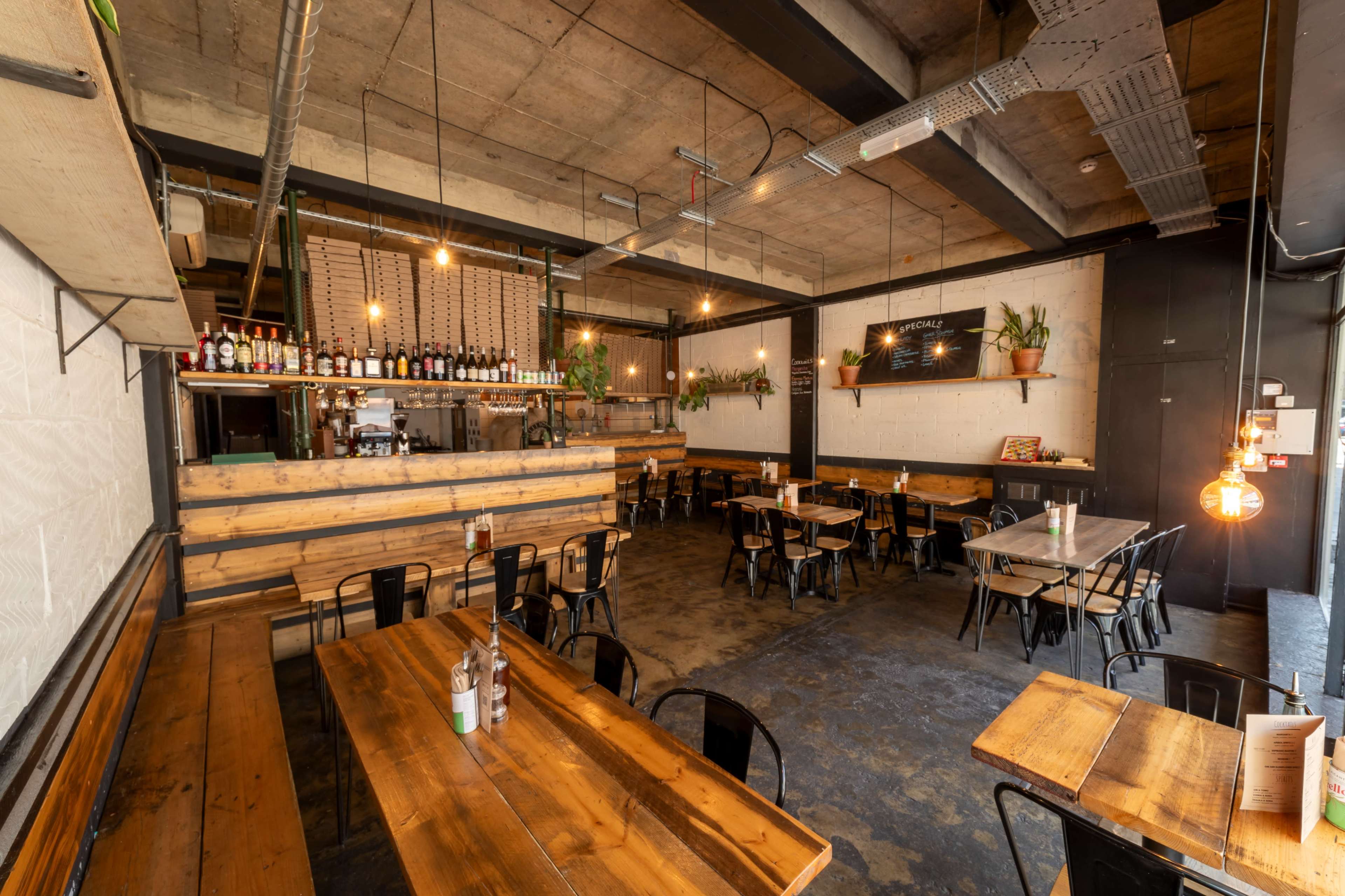 The image shows a modern restaurant interior with wooden tables and black metal chairs, featuring a bar area stocked with bottles and hanging light fixtures.