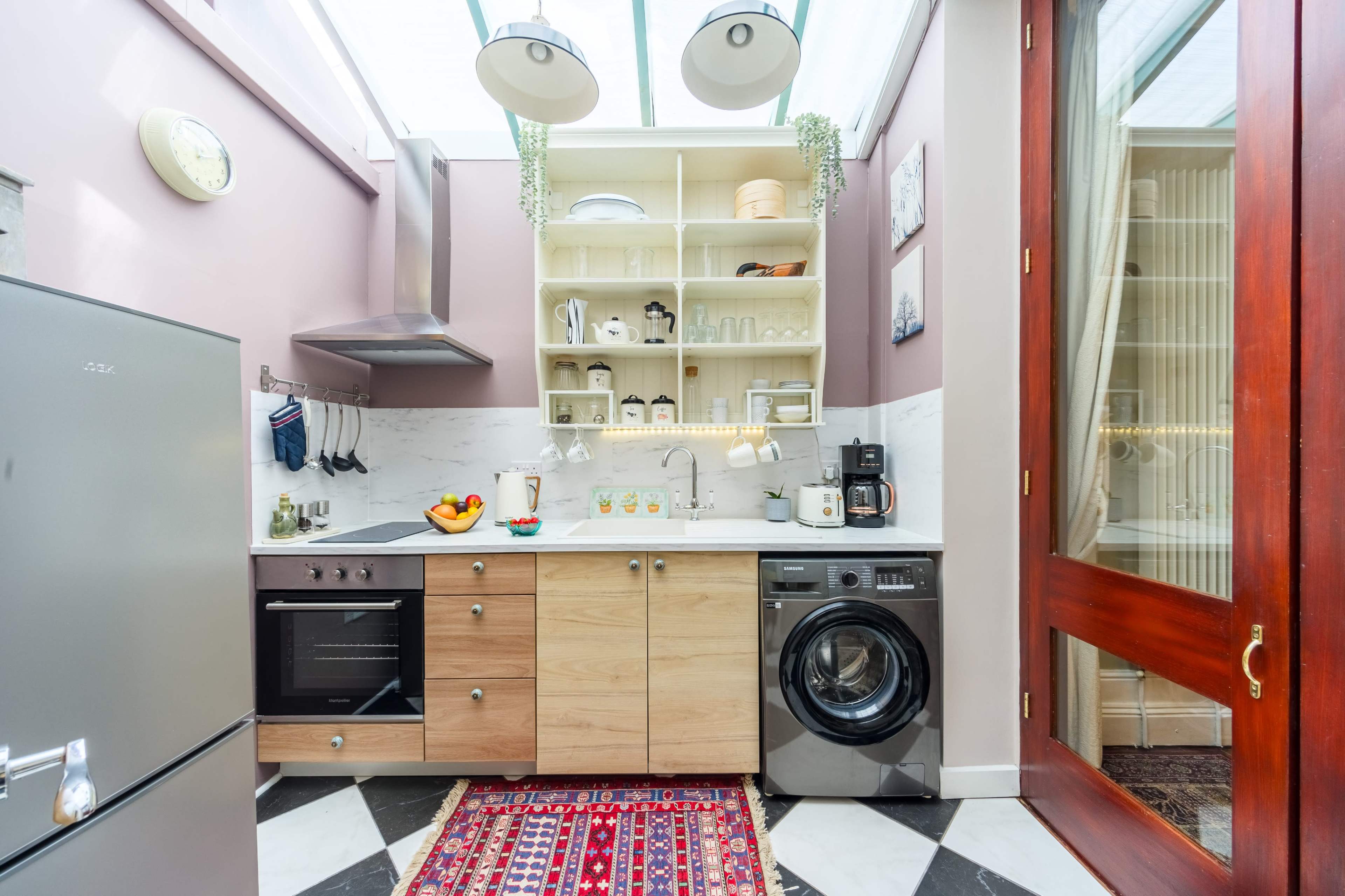 A modern kitchen features a stainless steel refrigerator, an oven, a washing machine, and open shelving displaying various dishes and decor against a backdrop of pink walls and marble countertops.