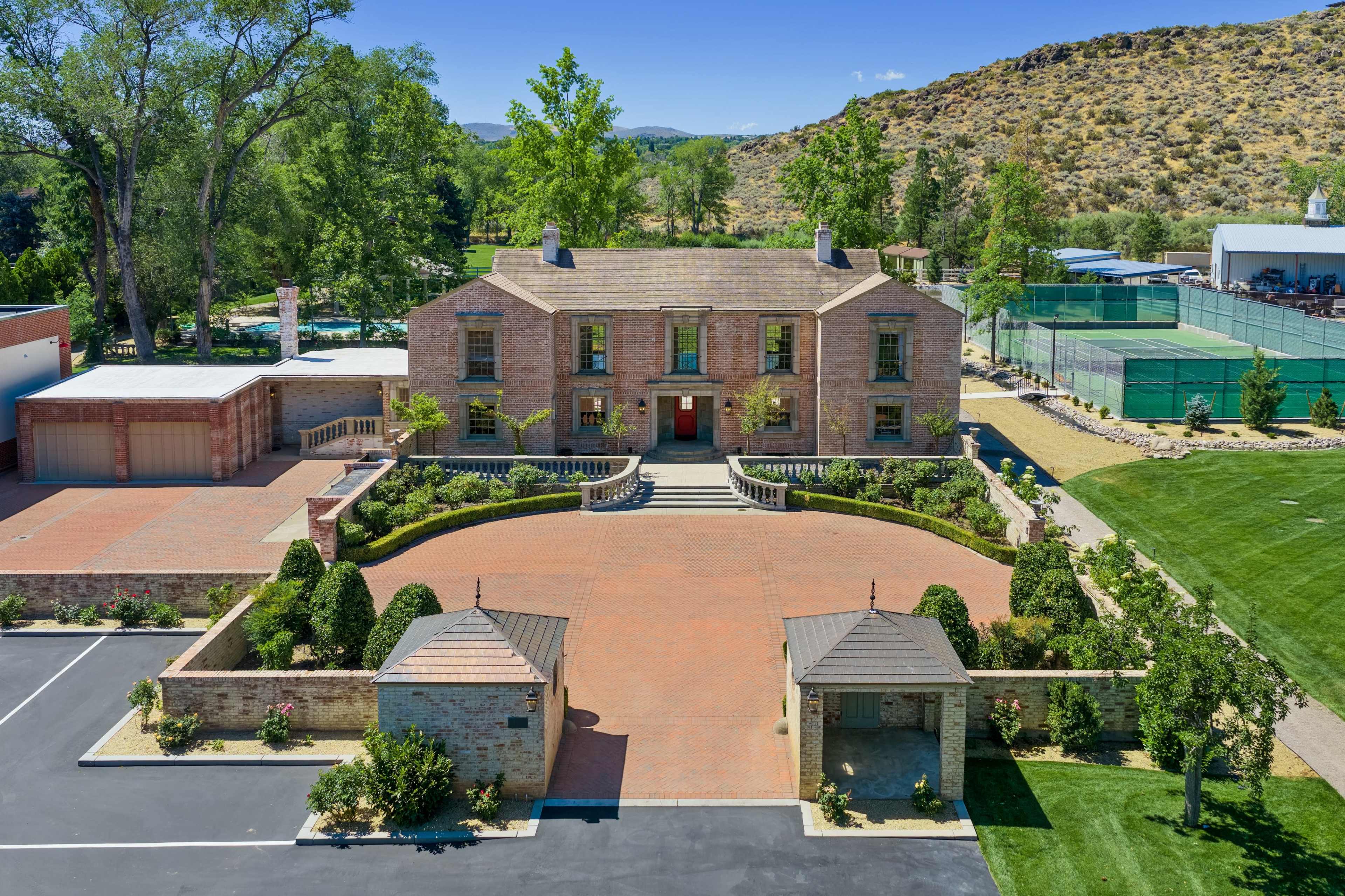 The image shows a large, two-story brick mansion with a landscaped driveway, surrounded by greenery and hills in the background.