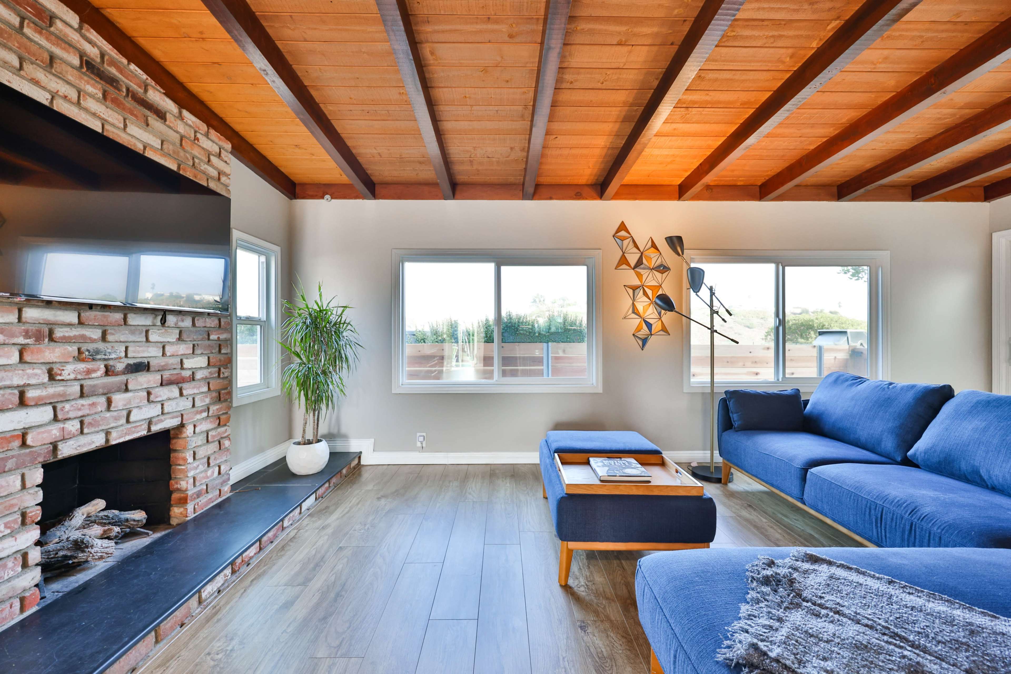 A living room features a brick fireplace, large windows that let in natural light, and a blue sectional sofa with a wooden coffee table.