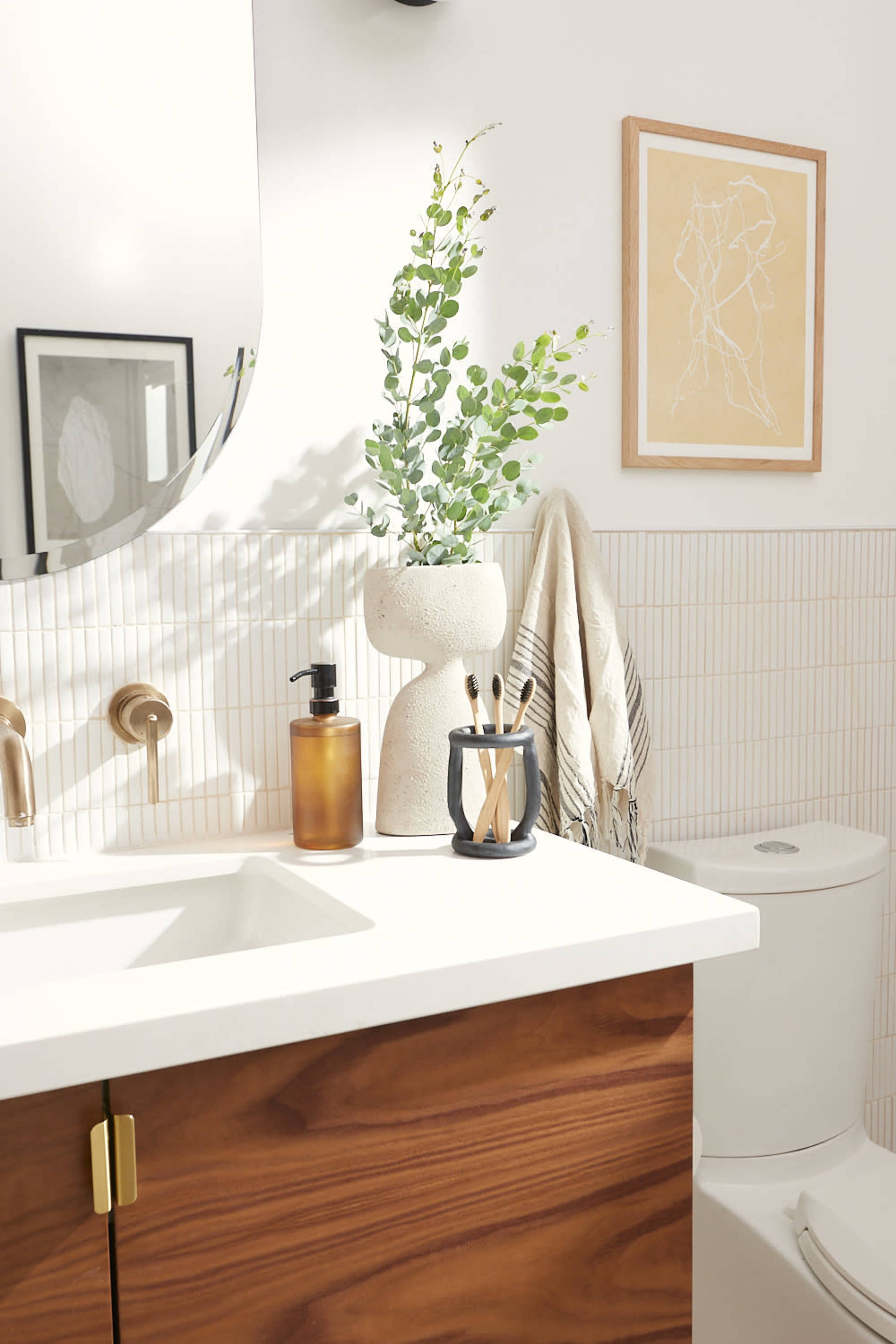 The image shows a modern bathroom with a sink, wooden cabinetry, a round mirror, potted greenery, and a tidy arrangement of toiletries.