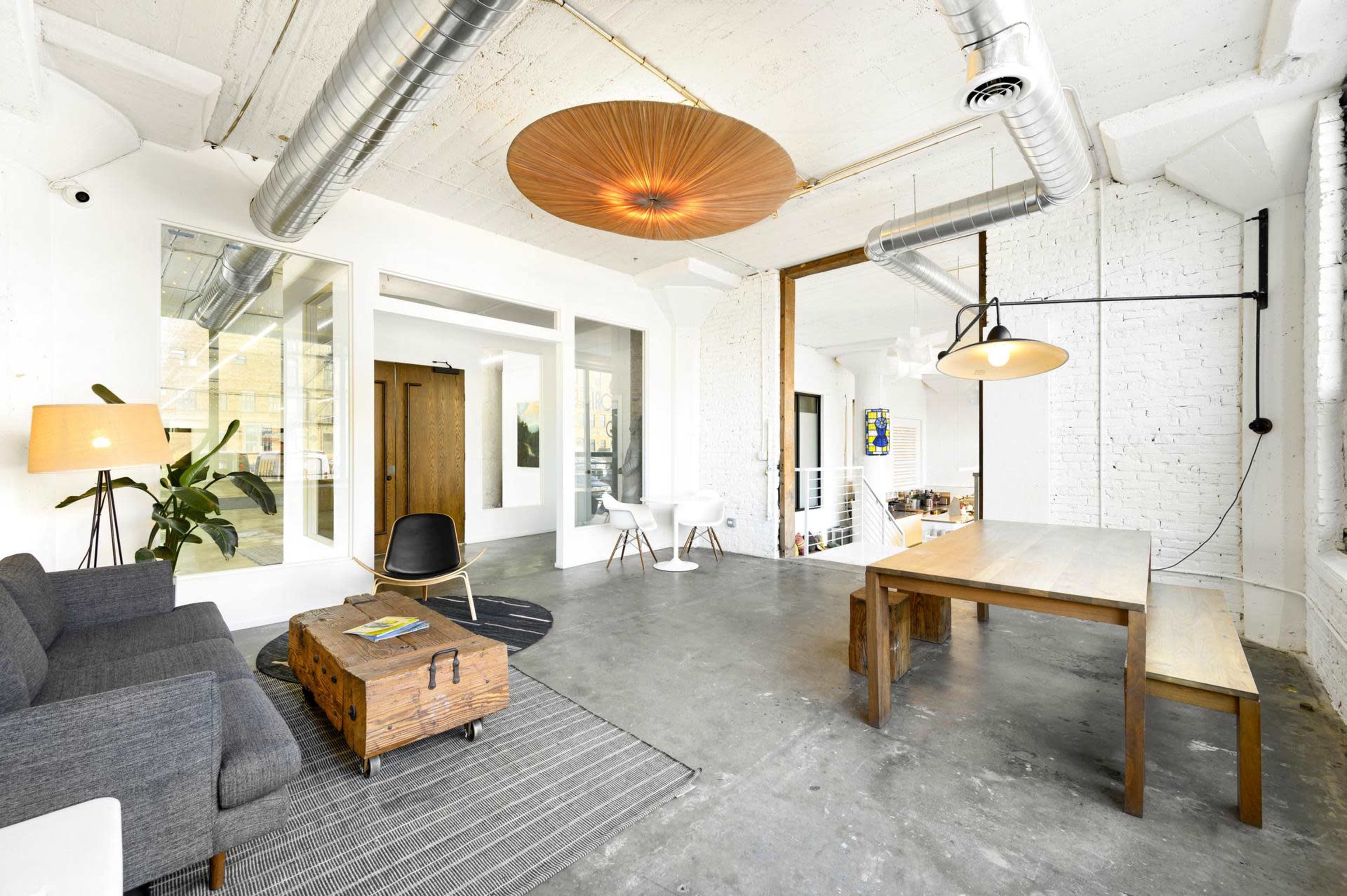 The image depicts a bright, modern open-concept space featuring a gray sofa, a wooden dining table, and various light fixtures, with a mix of industrial and minimalist design elements.