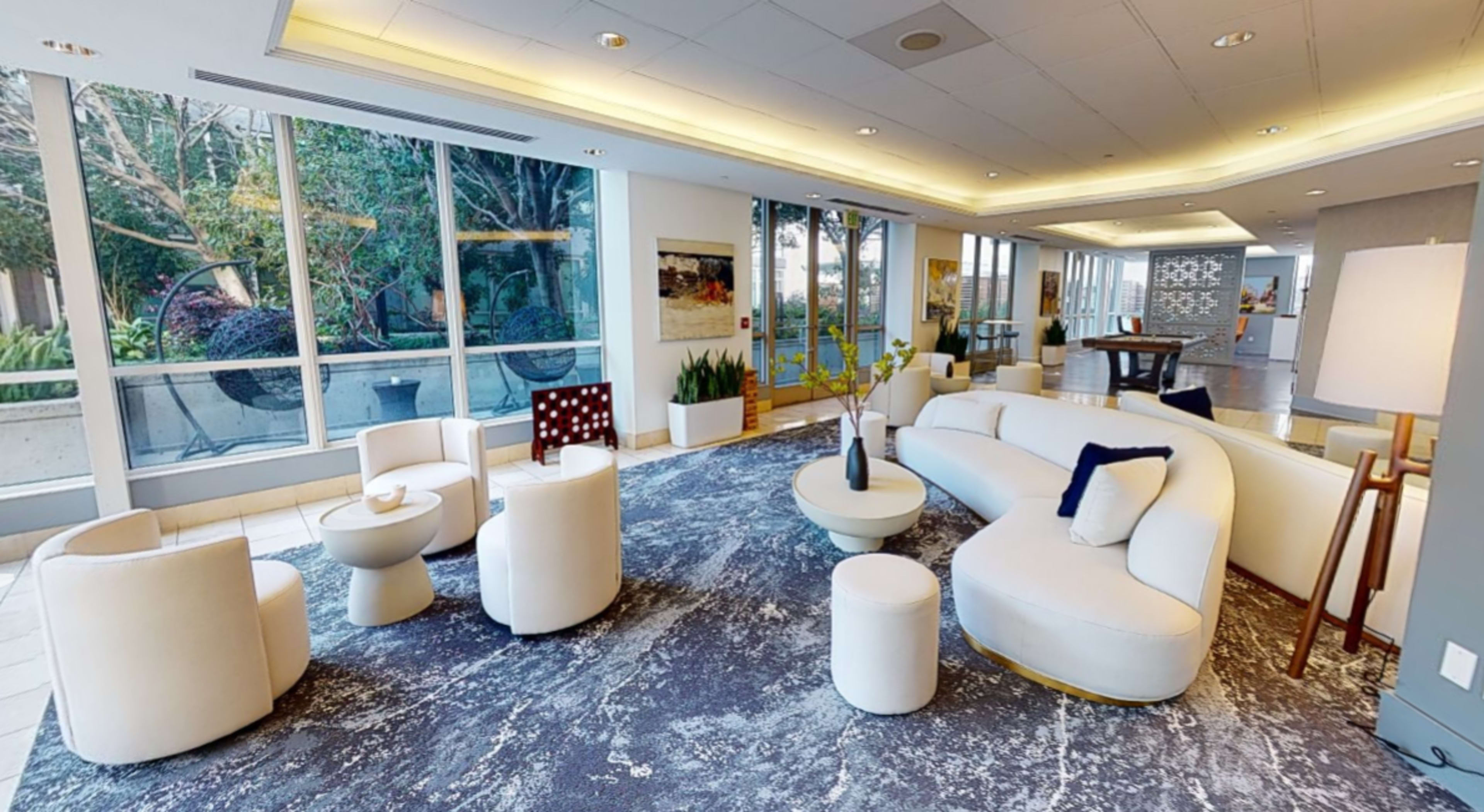 A modern lounge area with a white curved sofa, circular tables, and several chairs, adjacent to large windows offering views of greenery outside.