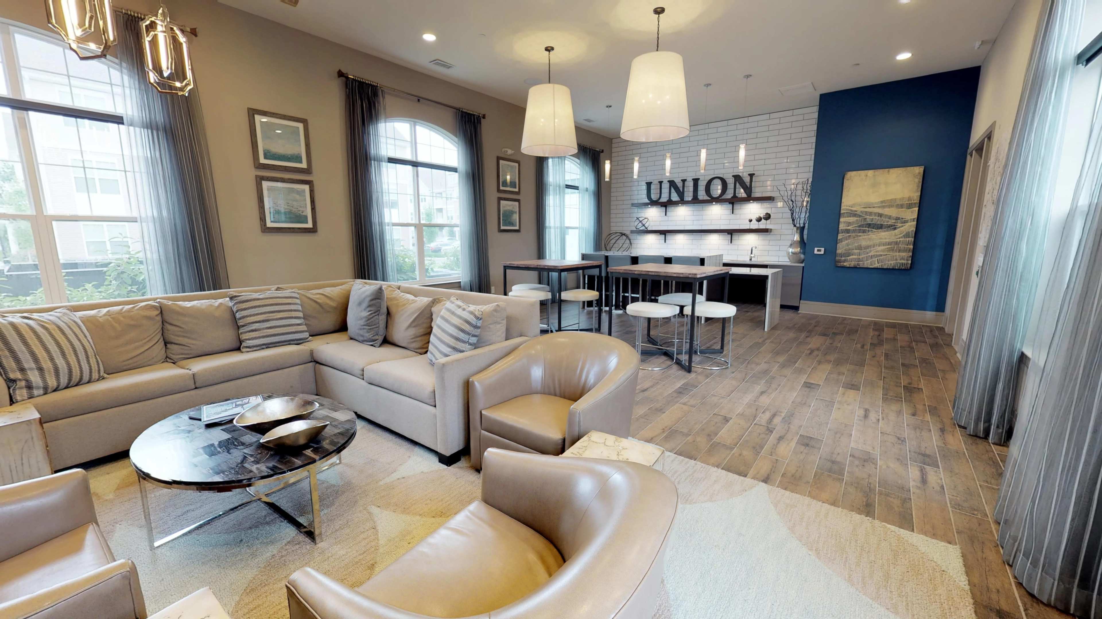 A modern lounge area with beige sofas, armchairs, a coffee table, and a kitchen bar in the background featuring the word "UNION" displayed on the wall.