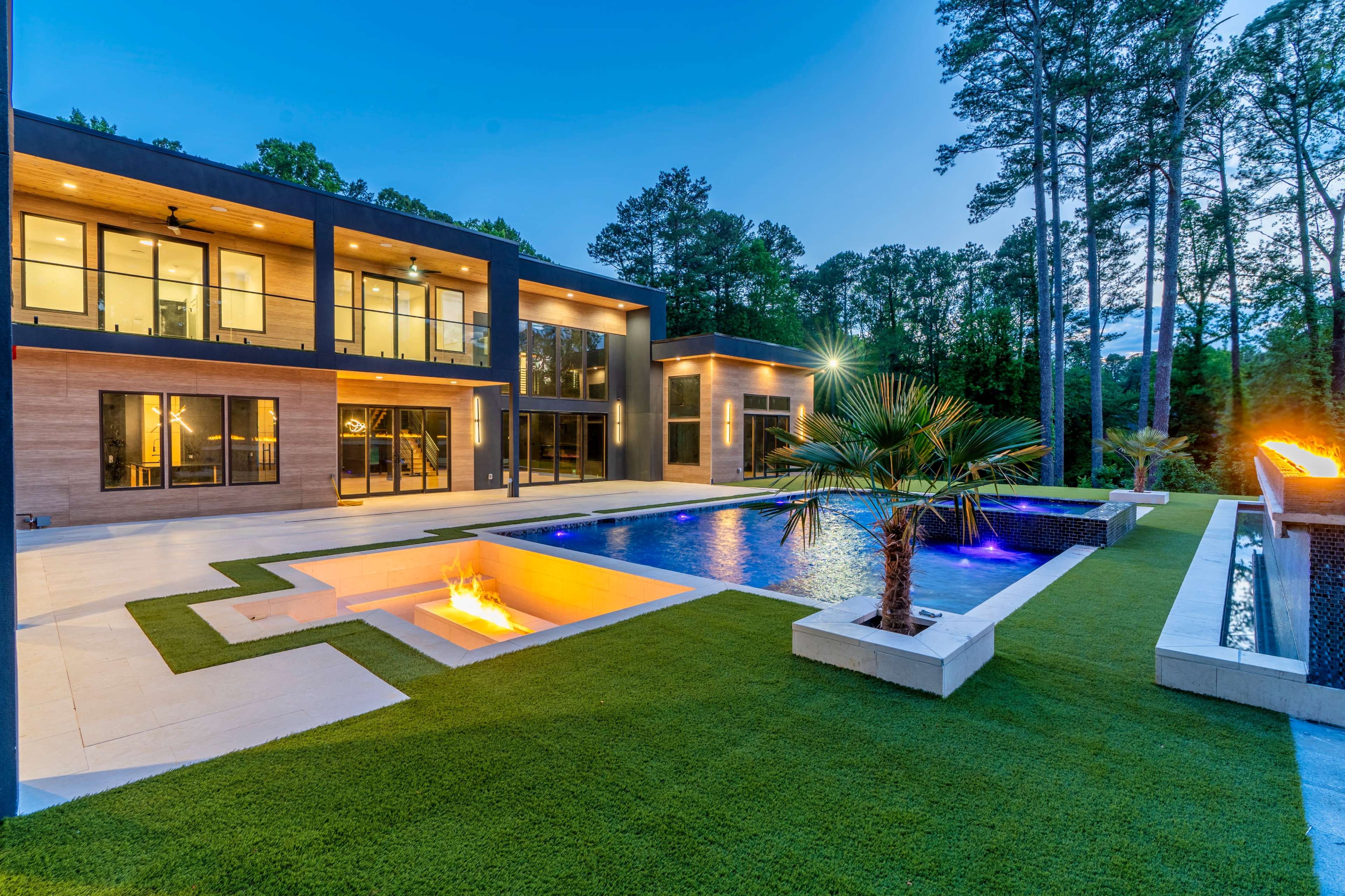 A modern house with large windows overlooks a pool surrounded by grass and palm trees, illuminated by lights in the evening.