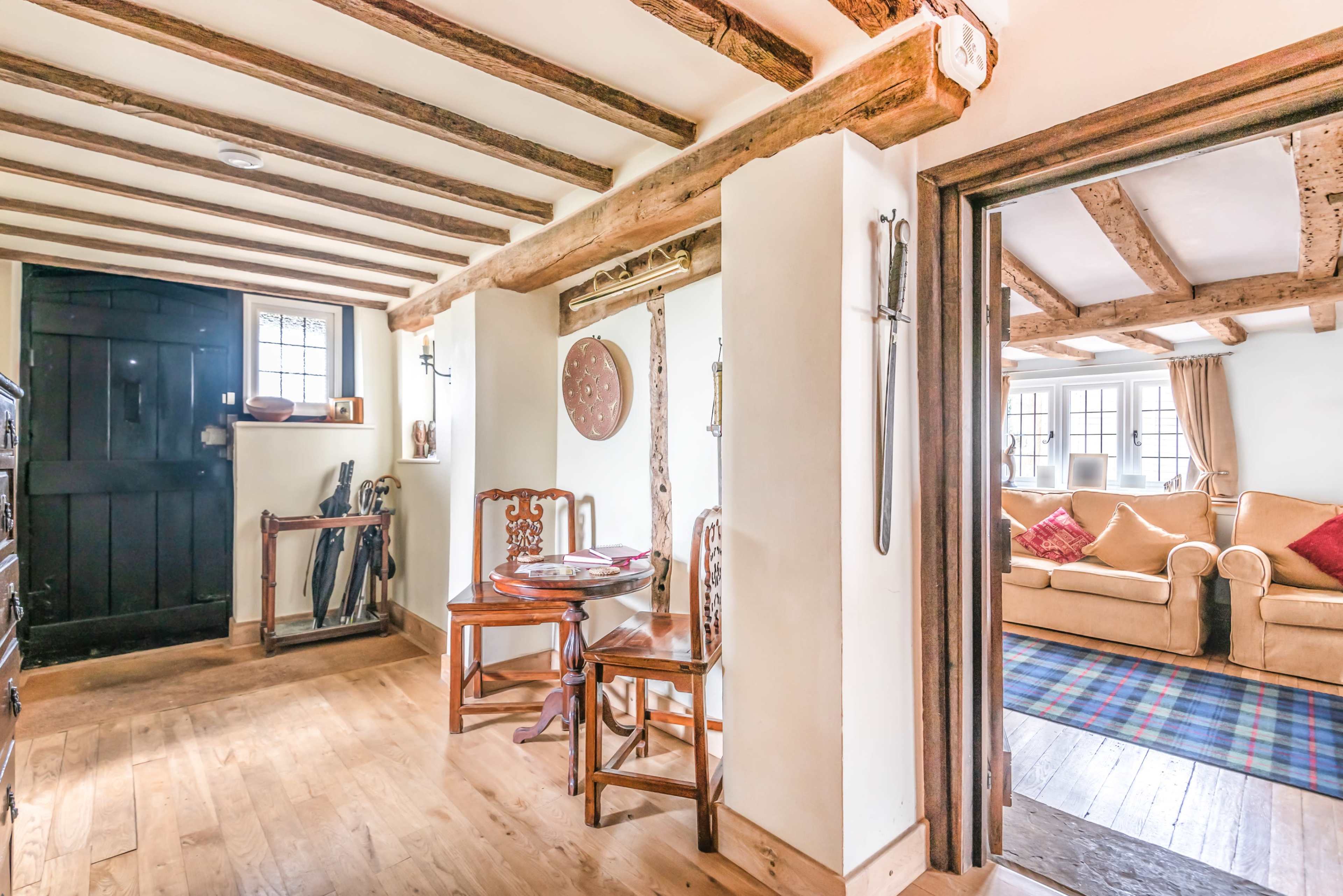 A rustic interior hallway with exposed wooden beams, a black door, and a small dining set, leading to a living room with a couch and plaid rug.