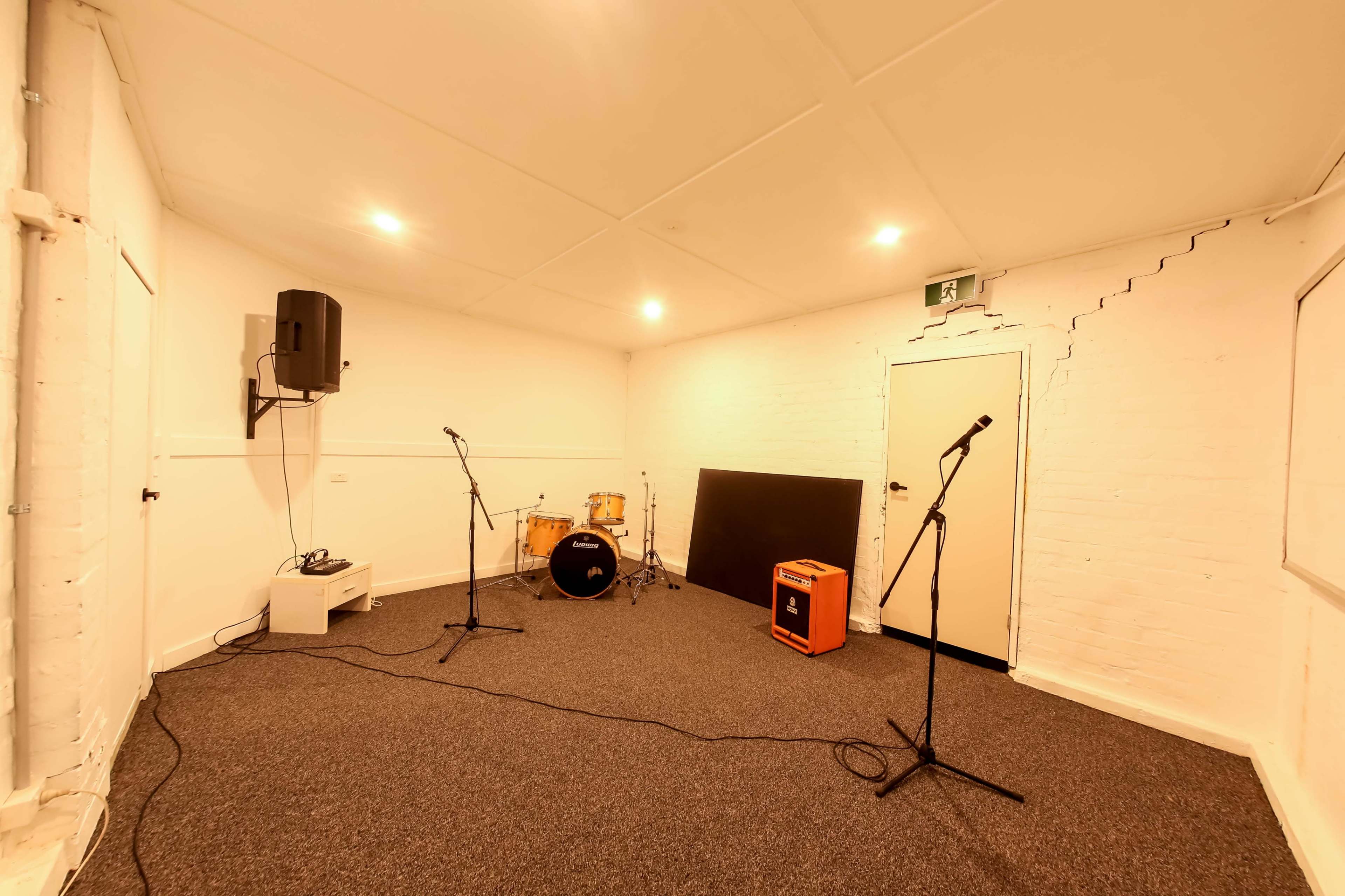 The image shows a small, empty room designed for music practice, featuring a drum set, microphones on stands, and an amplifier.
