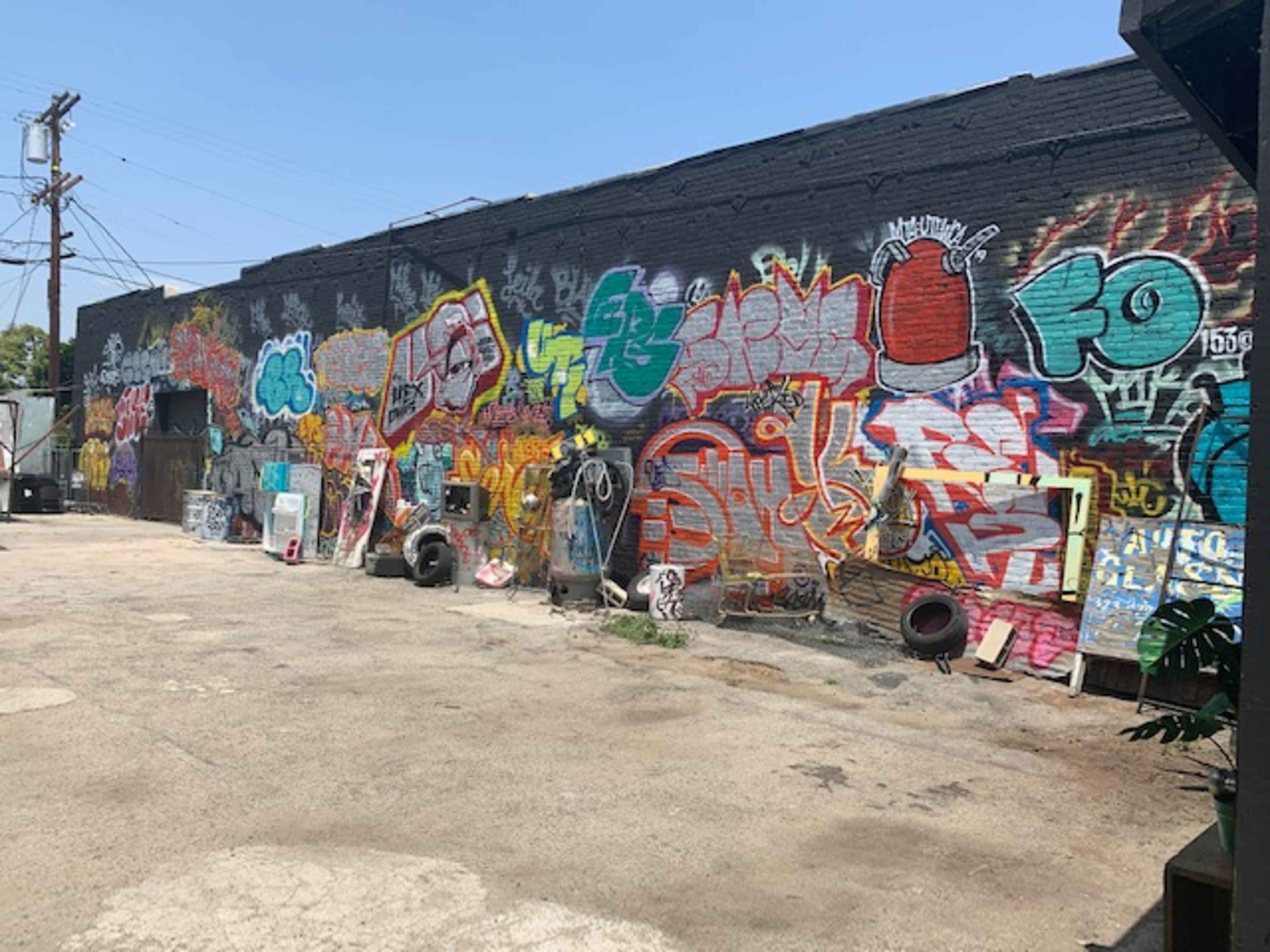 A dusty alleyway with a wall covered in colorful graffiti and discarded objects scattered along the ground.