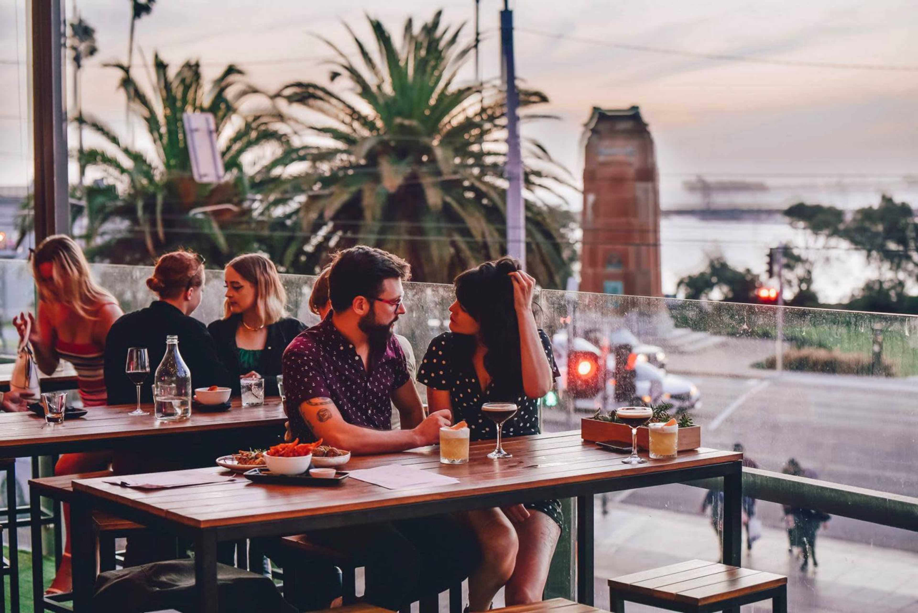 A couple sits at a table on a balcony of a restaurant, facing each other with drinks in front, while palm trees and a sunset are visible in the background.