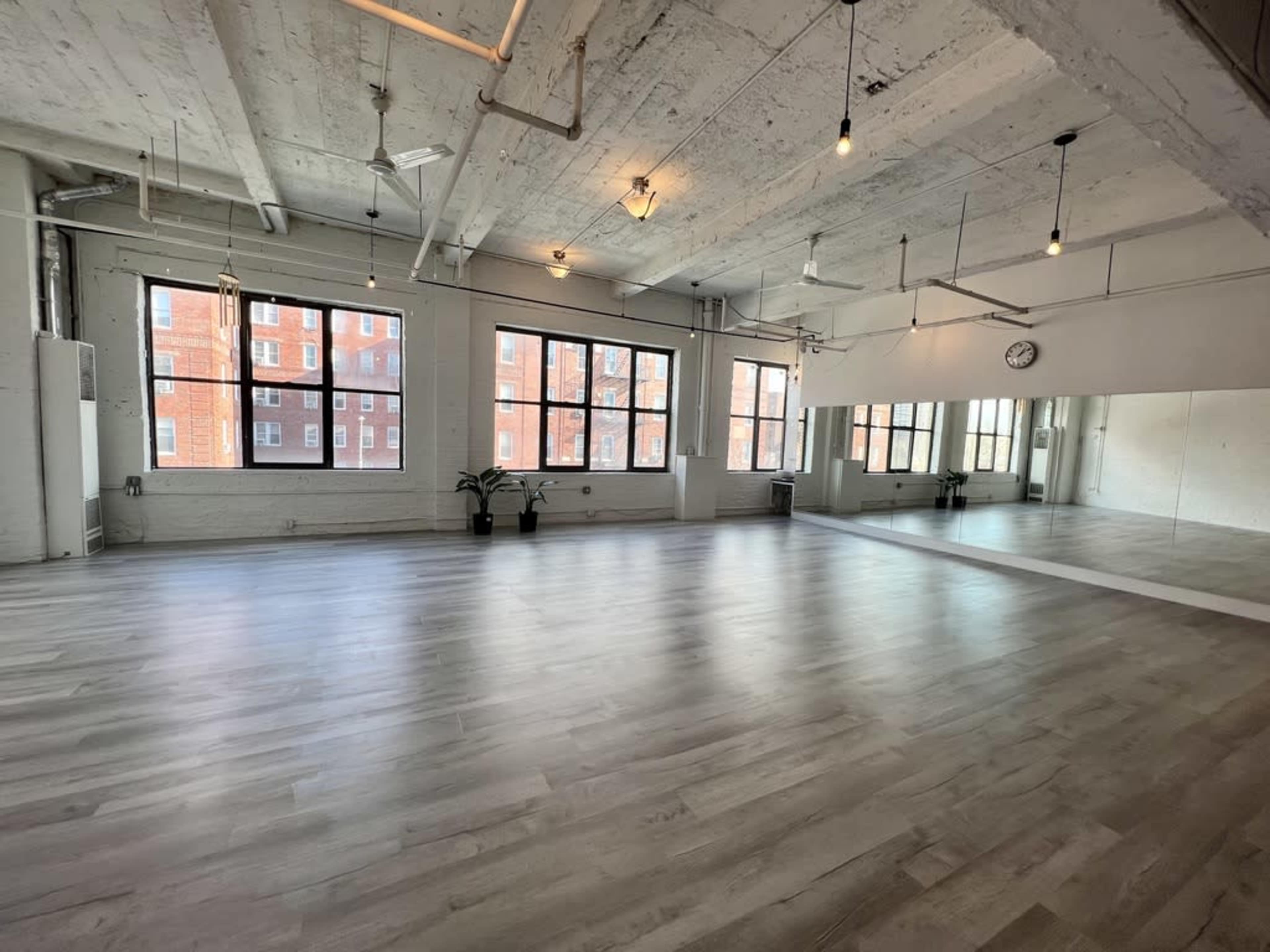 The image shows a spacious, well-lit studio with large windows and polished wooden flooring, featuring a mirror along one wall.
