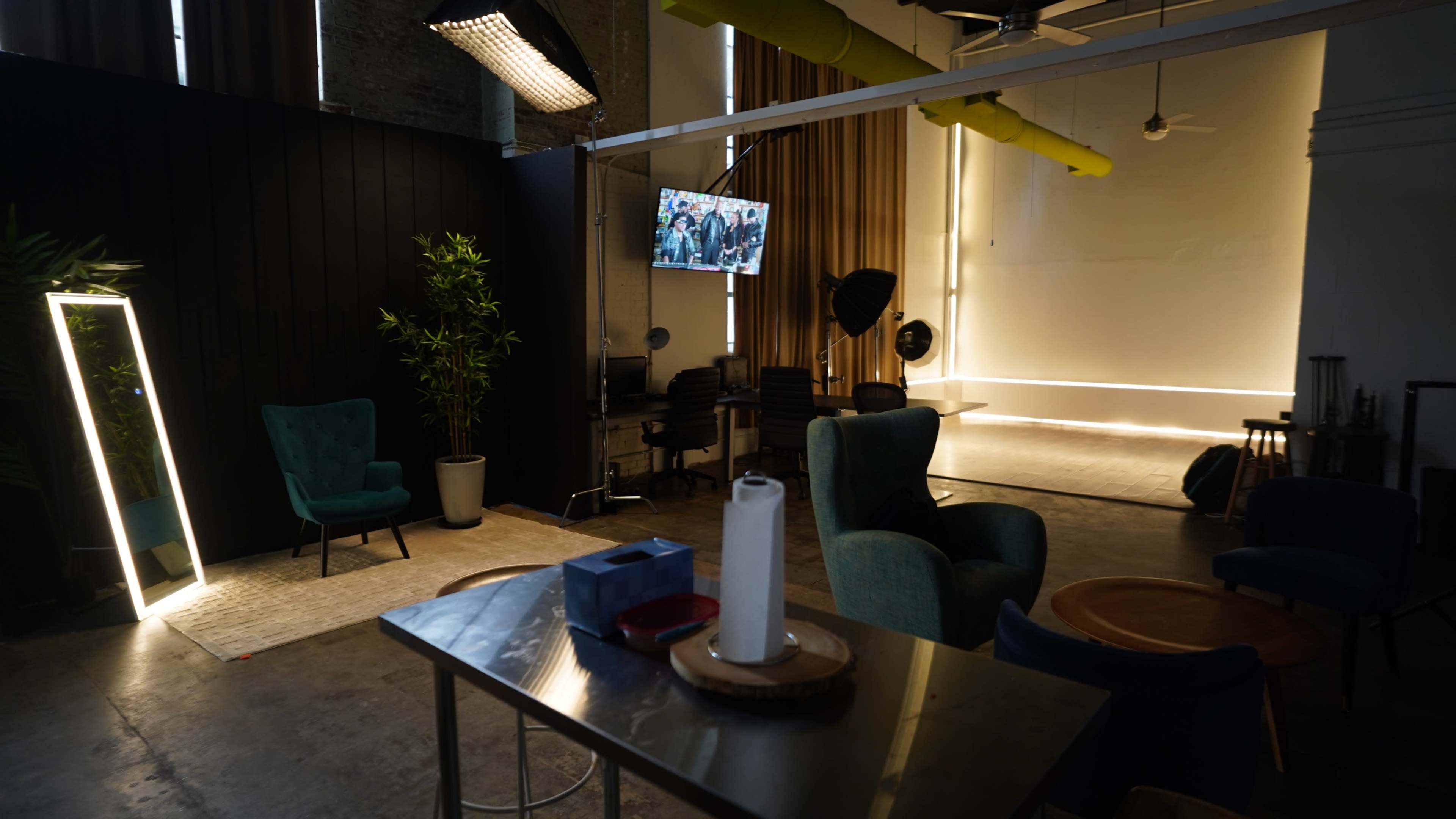 A modern studio space features green armchairs, a tall plant, a metal table, and a wall-mounted TV displaying a scene, with bright lighting and a neutral backdrop.