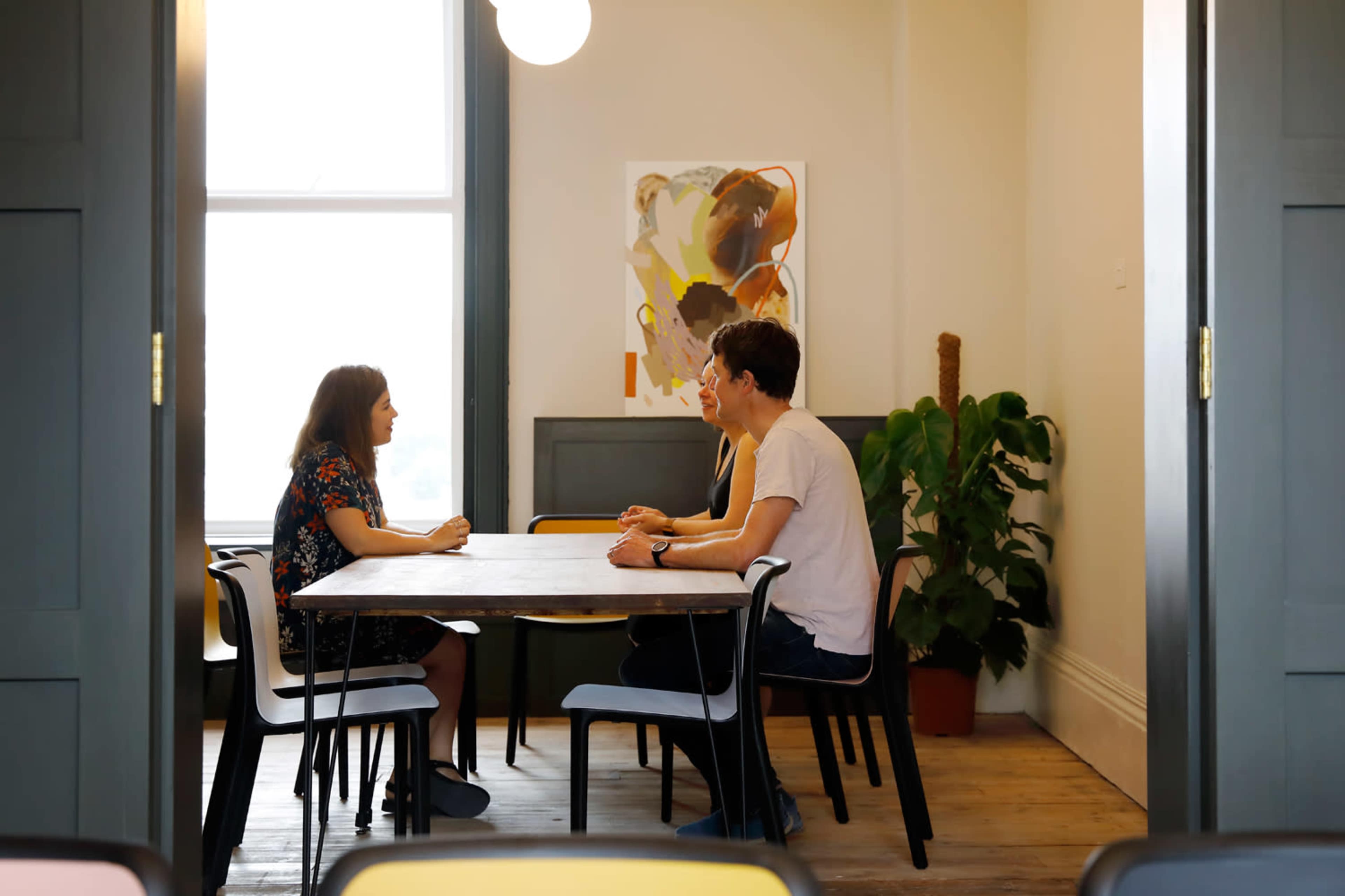 Three people are sitting at a table in a bright room, engaged in conversation, with a large window and artwork on the wall.