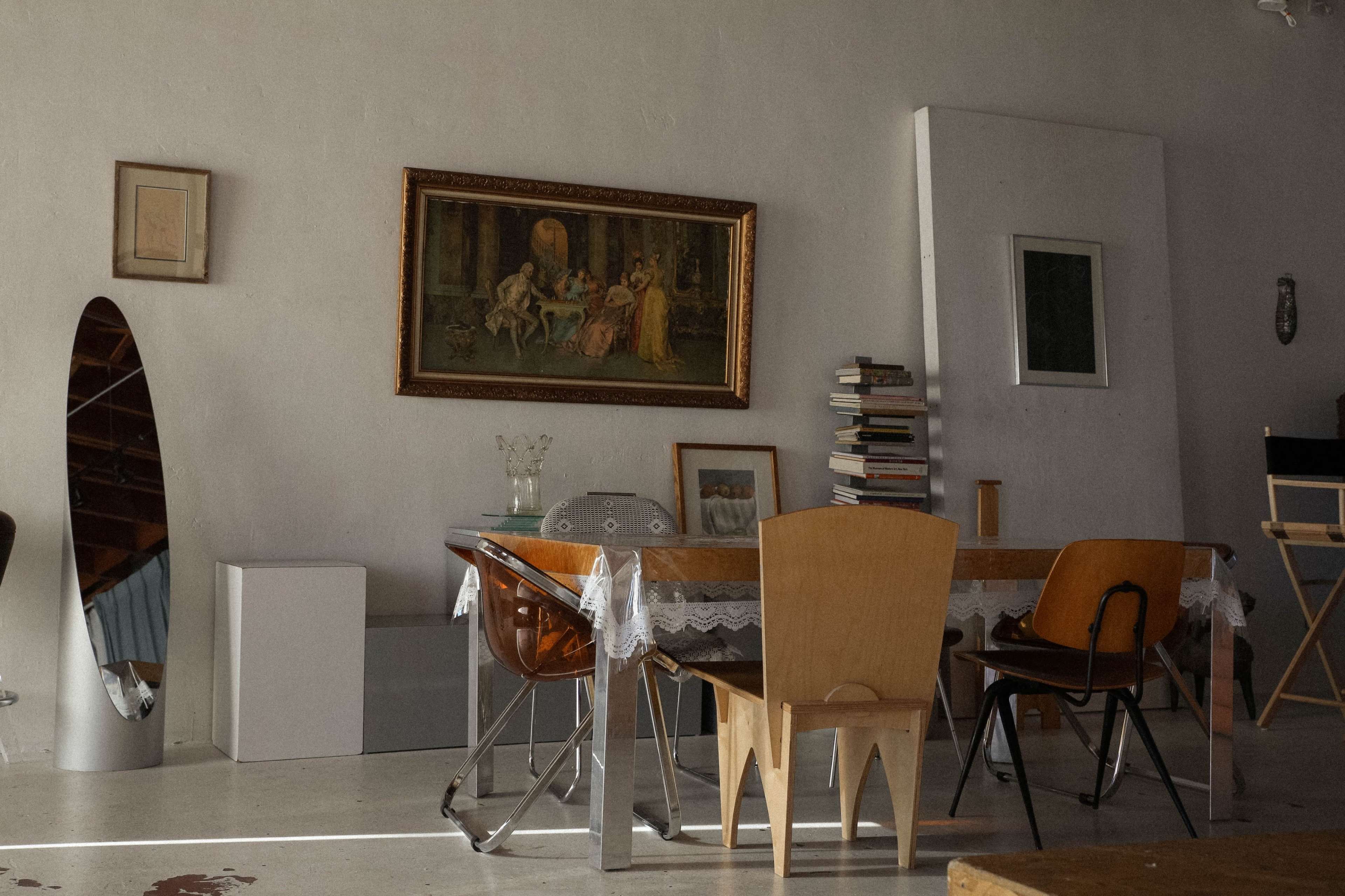 A modern, minimalist dining area features a wooden table surrounded by various styled chairs, with artwork on the walls and a large mirror reflecting the space.