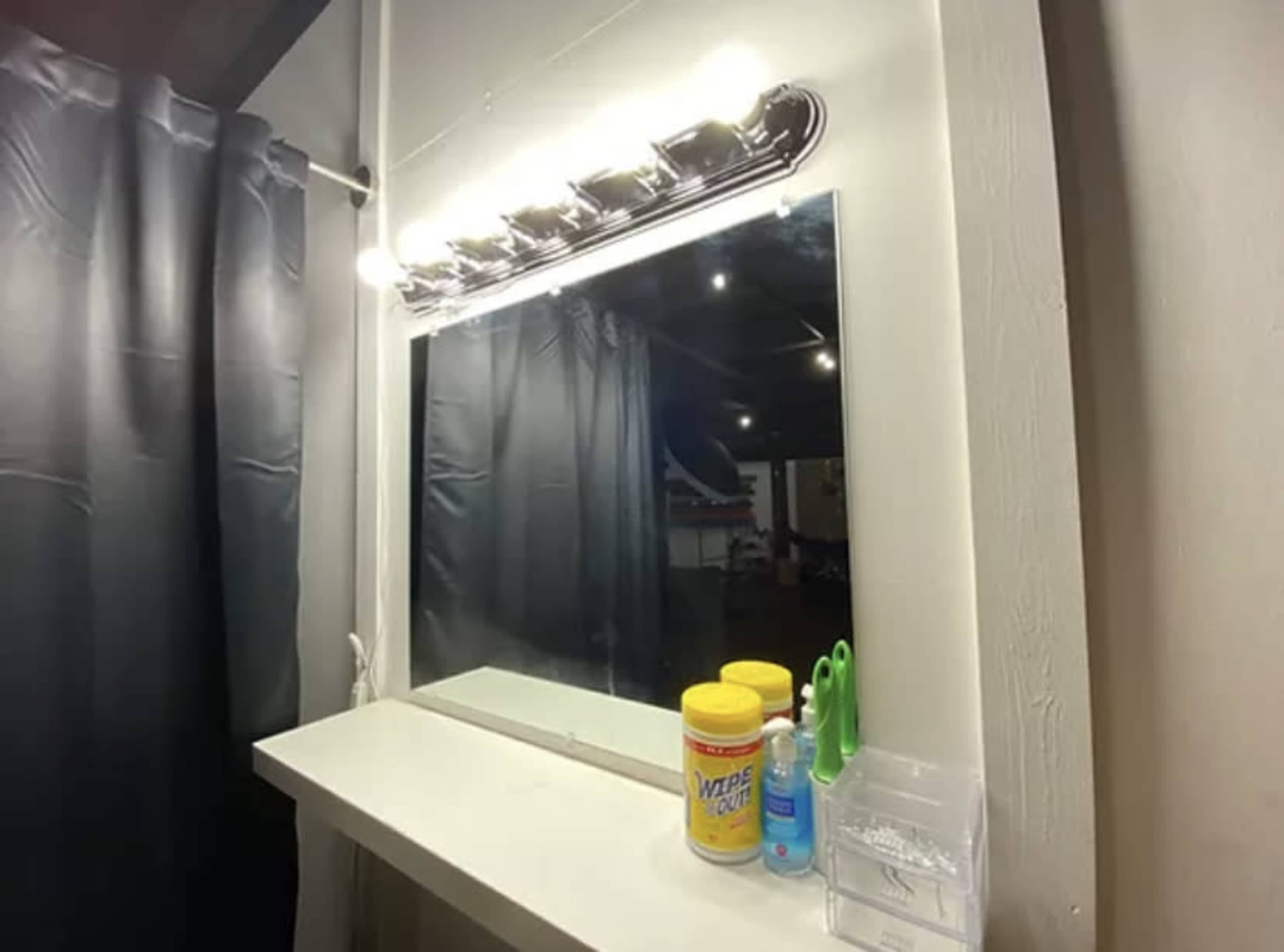 A bathroom vanity with a large mirror, bright light fixture above it, and cleaning supplies on the shelf below.