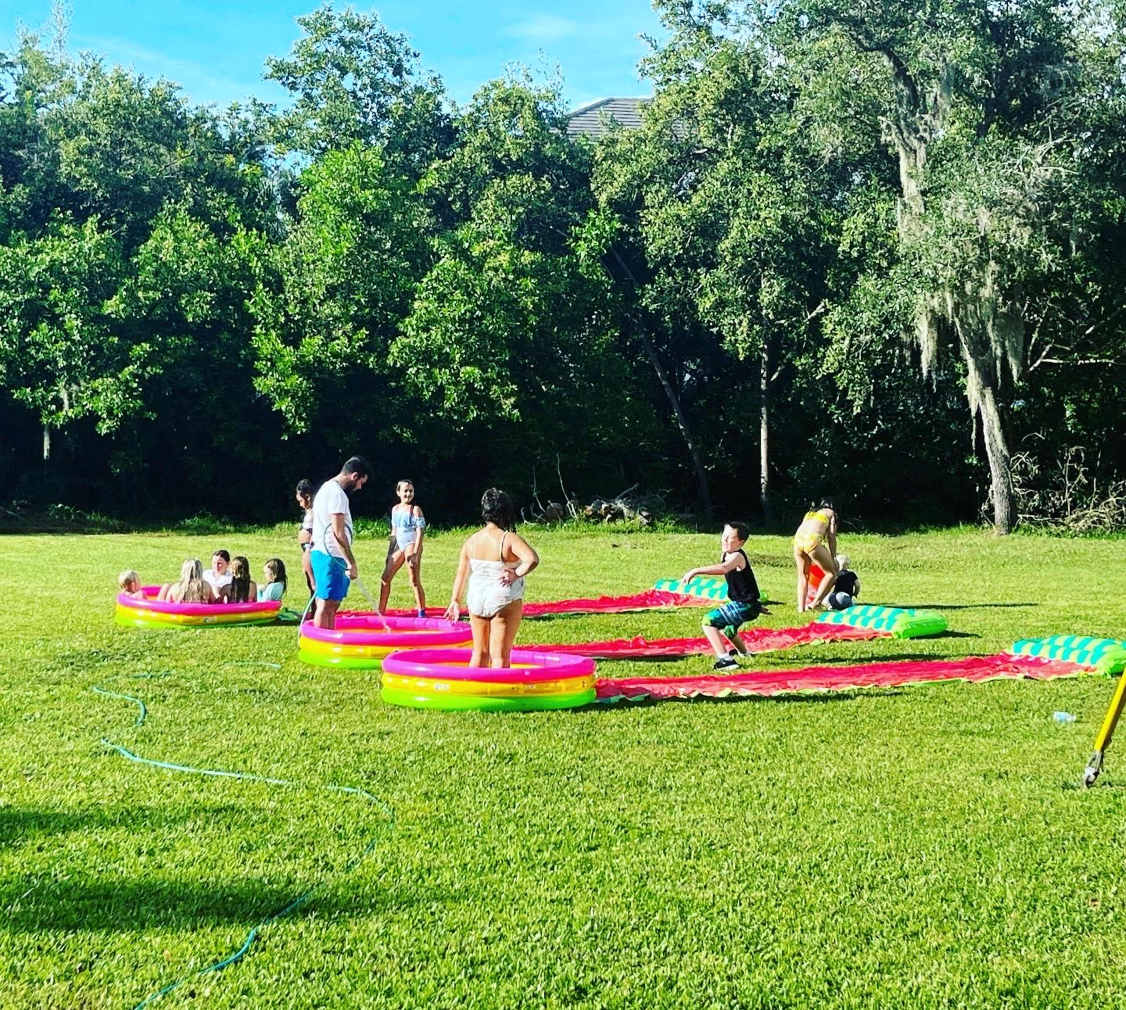 A group of people are playing on inflatable water slides set up on a grassy area surrounded by trees.